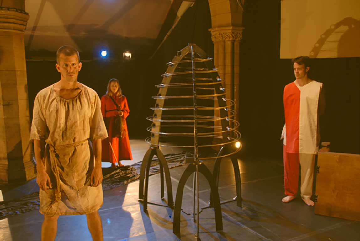 Three actors stand on a stage, in costume, surrounding a metal conical structure.