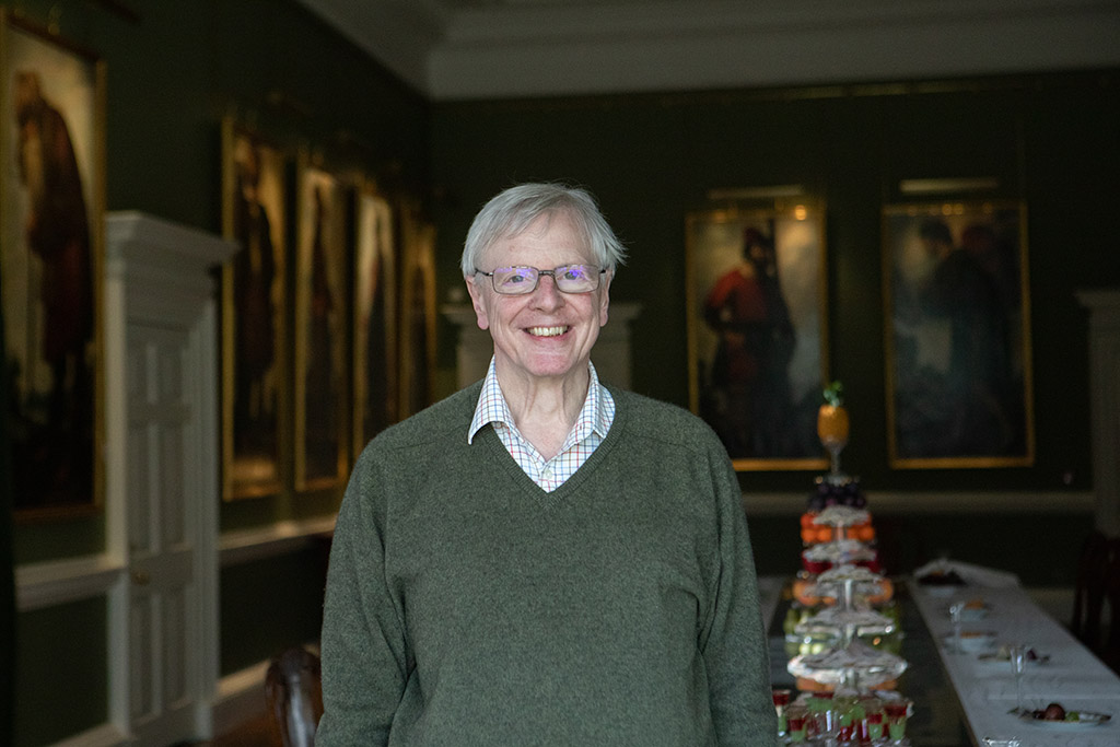 A man stands in a formal dining room that has traditional paintings on the walls