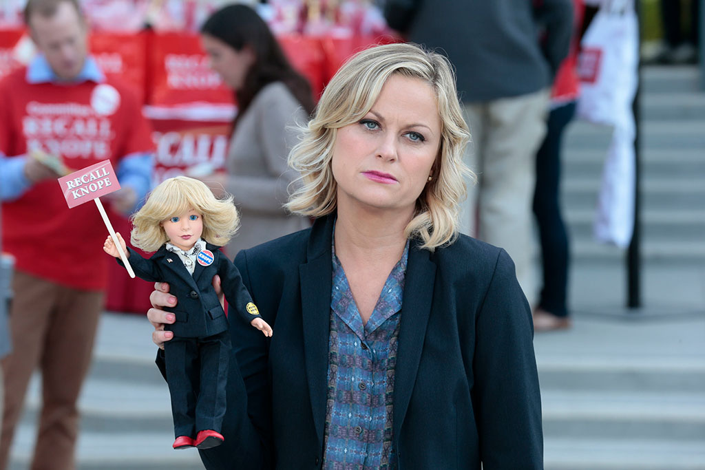 A deflated looking woman stands aside from a protest rally, holding a small doll of herself that reads Recall Knope'.