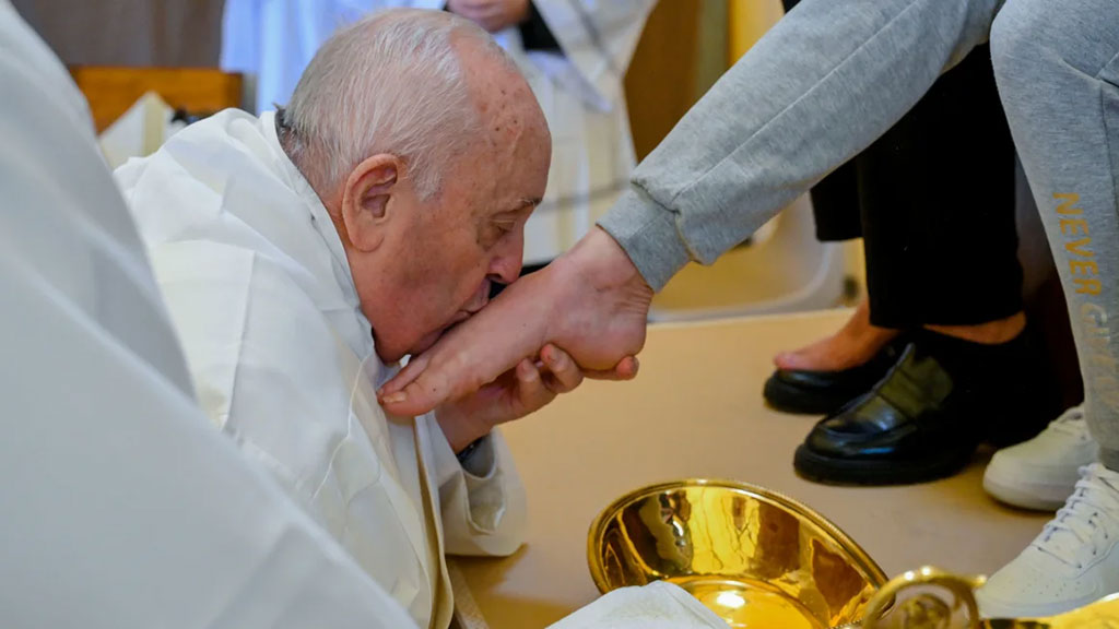 The Pope, wearing white, kneels, crades a bare foot, and kisses it.