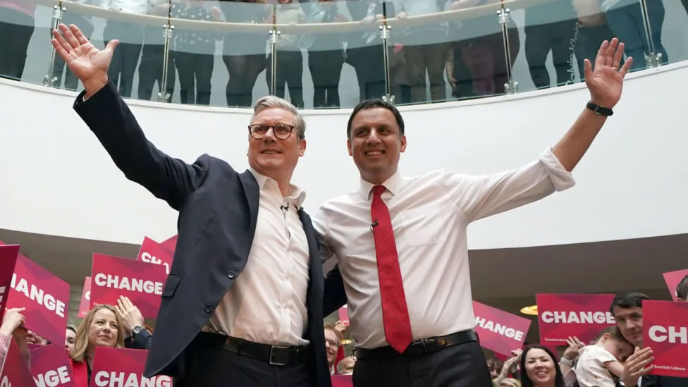 Two politicians, stand with one arm around each other and the other arms raised.