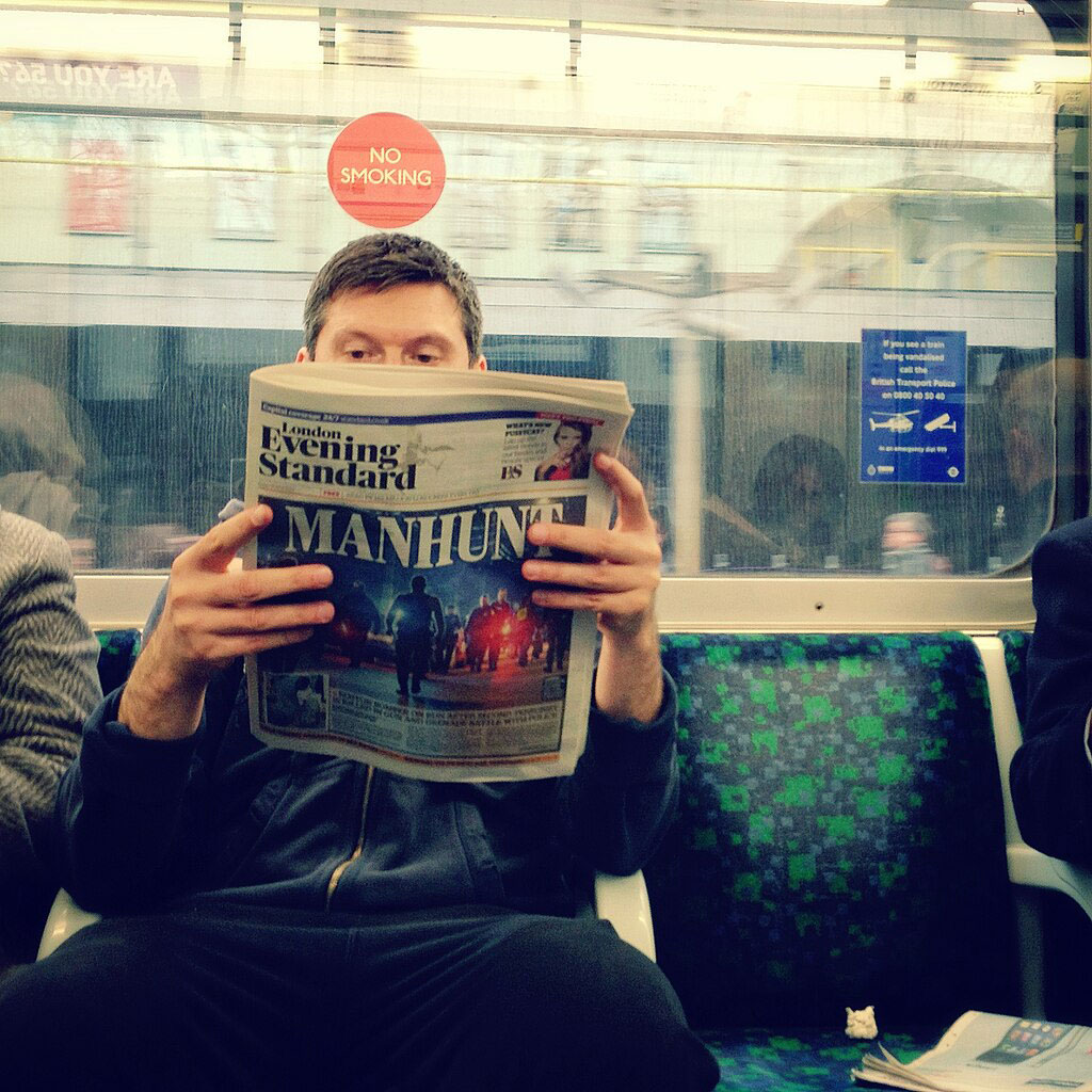 Seat on an underground train carraige, a passenger holds and reads a newspaper.