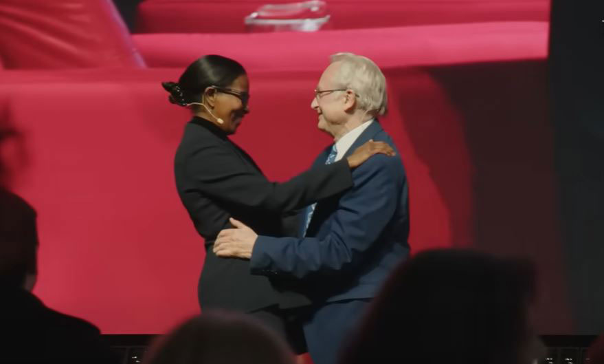 A man and woman speaker on a stage greet and embrace each other.