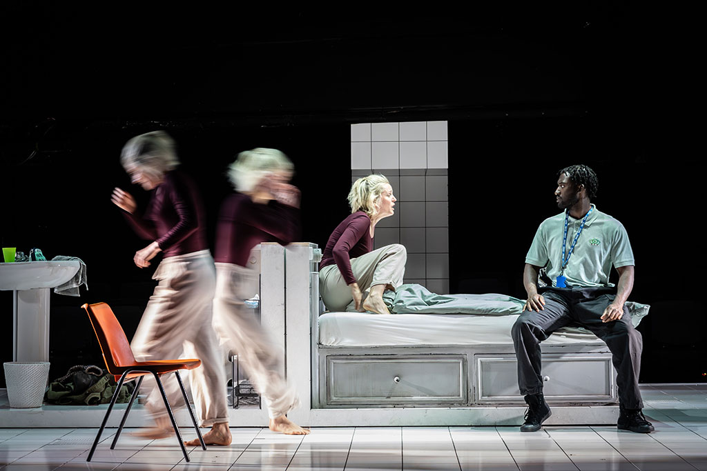 on a stage a woman kneels on a bed amid frantic action around her.