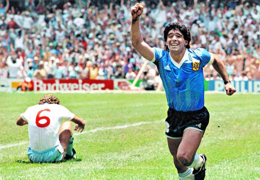 Maradona runs in celebration, holding a hand aloft as an England player sits dejected on the ground.