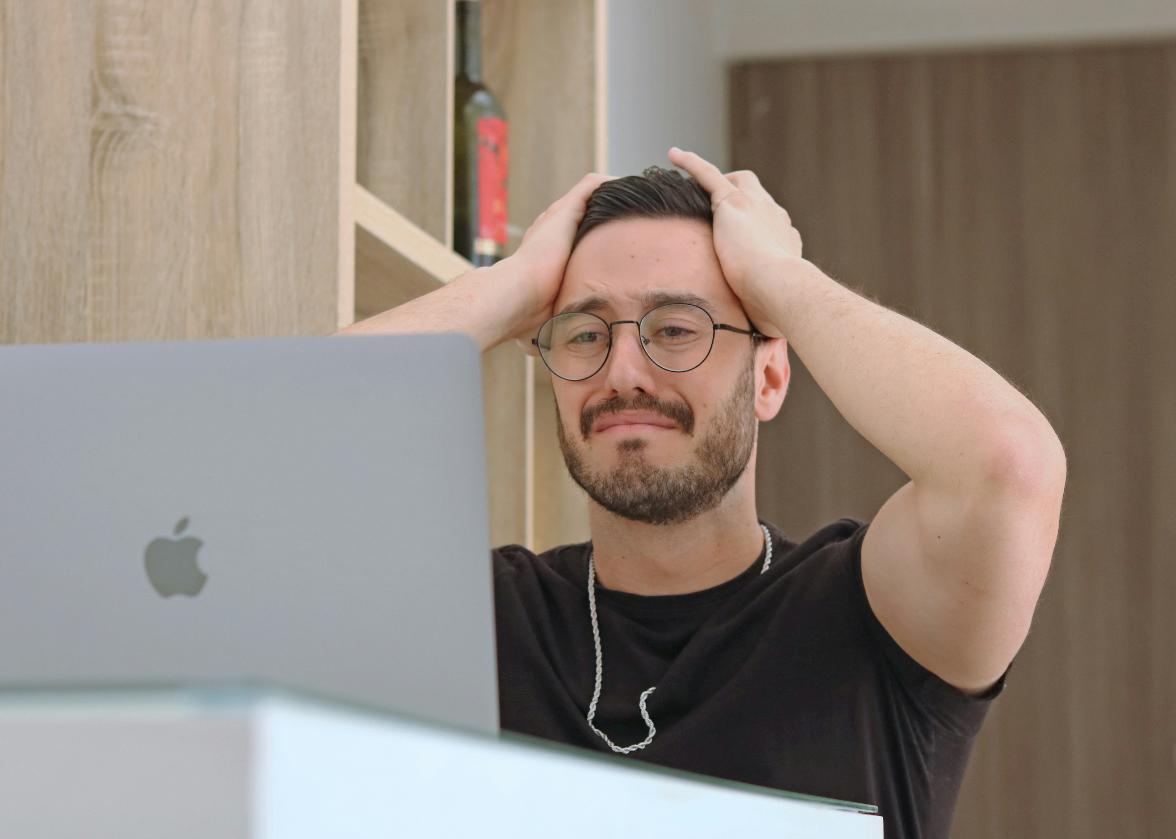 A man staring at a laptop grimmaces and holds his hands to his head.