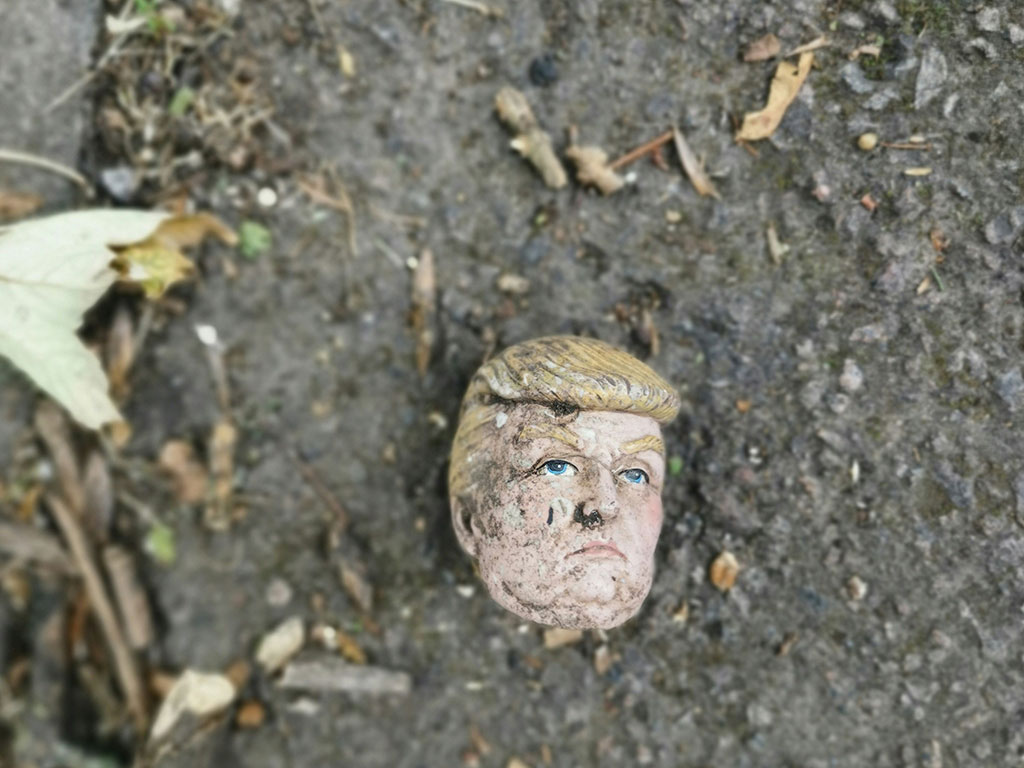 A severed doll head, resembling Donald Trump, lies on dirty ground.