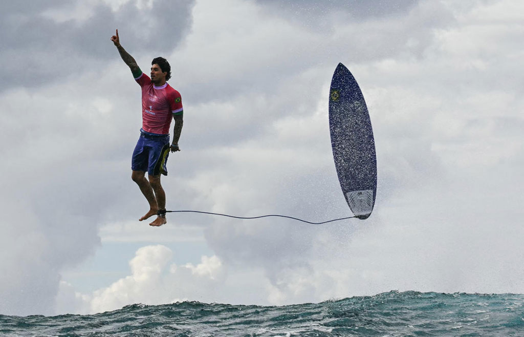 A surfer stands in the air, above a wave, with his board beside him.