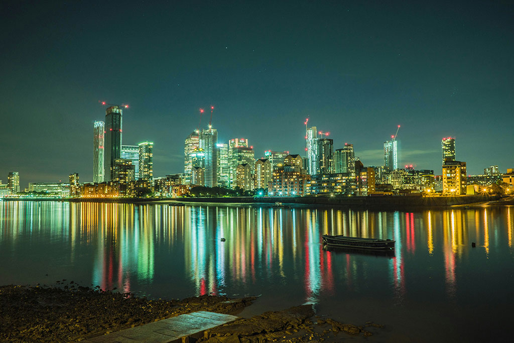 Skyscrapers loom under a dark sky and are reflected in a river in the foreground