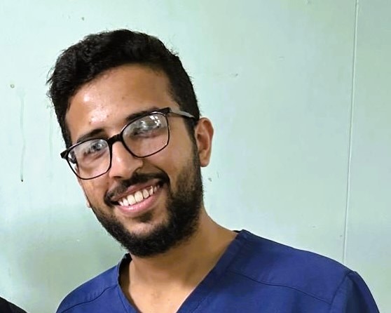 A young doctor wearing scrubs smiles.