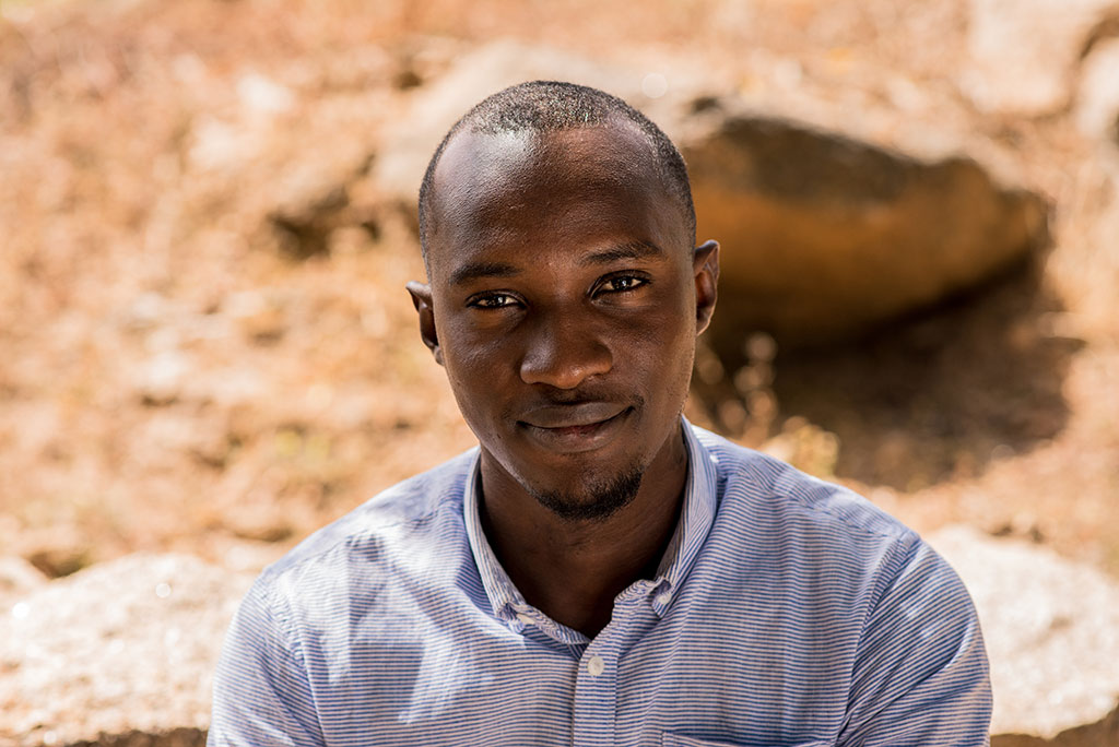 A Nigerian man looks up towards the camera, behind him is dusty ground