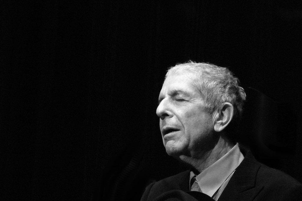 A black and white photo shows, singer Leonard Cohen to the right hand side, eyes closed and head inclined slightly upwards.