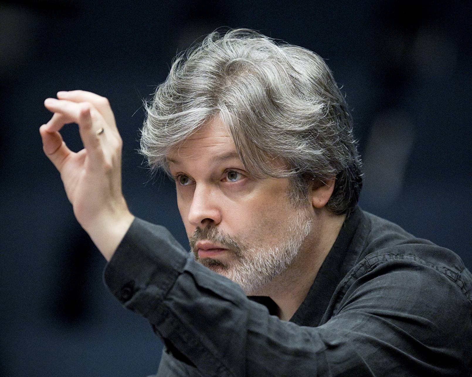 A conductor leans in toward an unseen orchestra with a raised hand.