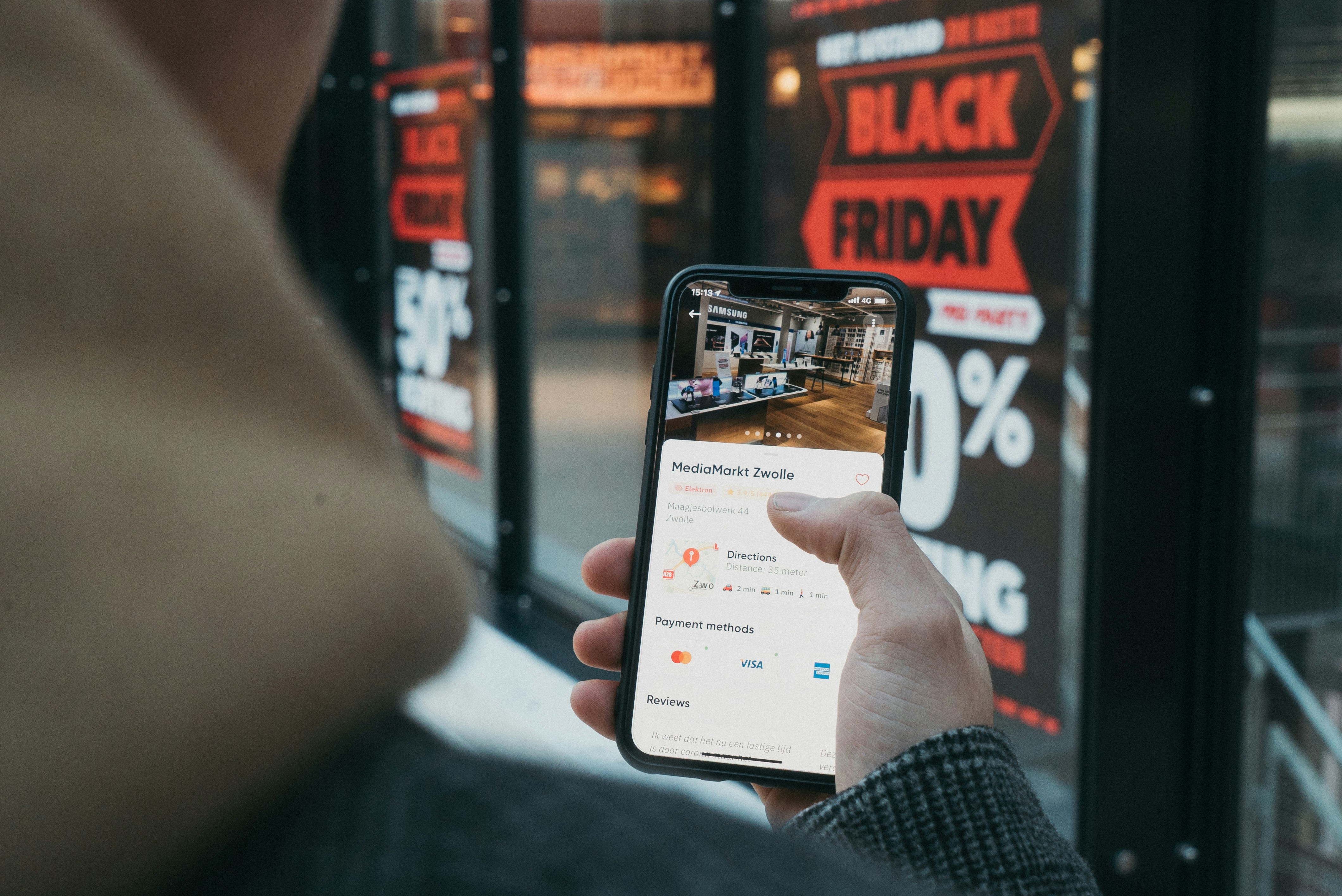 A phone screen is held up, showing a shopping page, behind a window displays a Black Friday Sale banner.