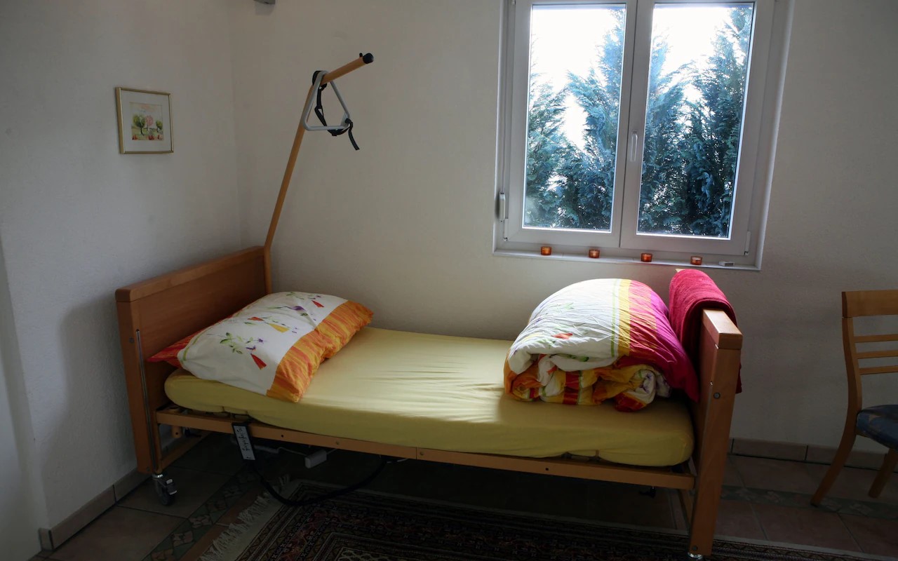 A single bed, wiith an unmade colourful duvet stands in the corner of a room. A hoist reaches over it from the corner. 