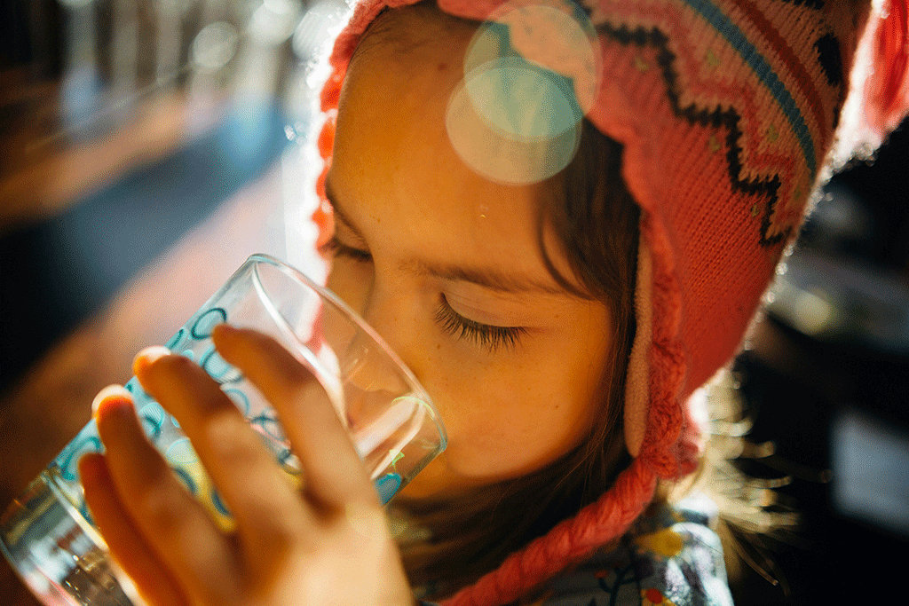 A child wearing a wool hat holds a glass and drink water from it.