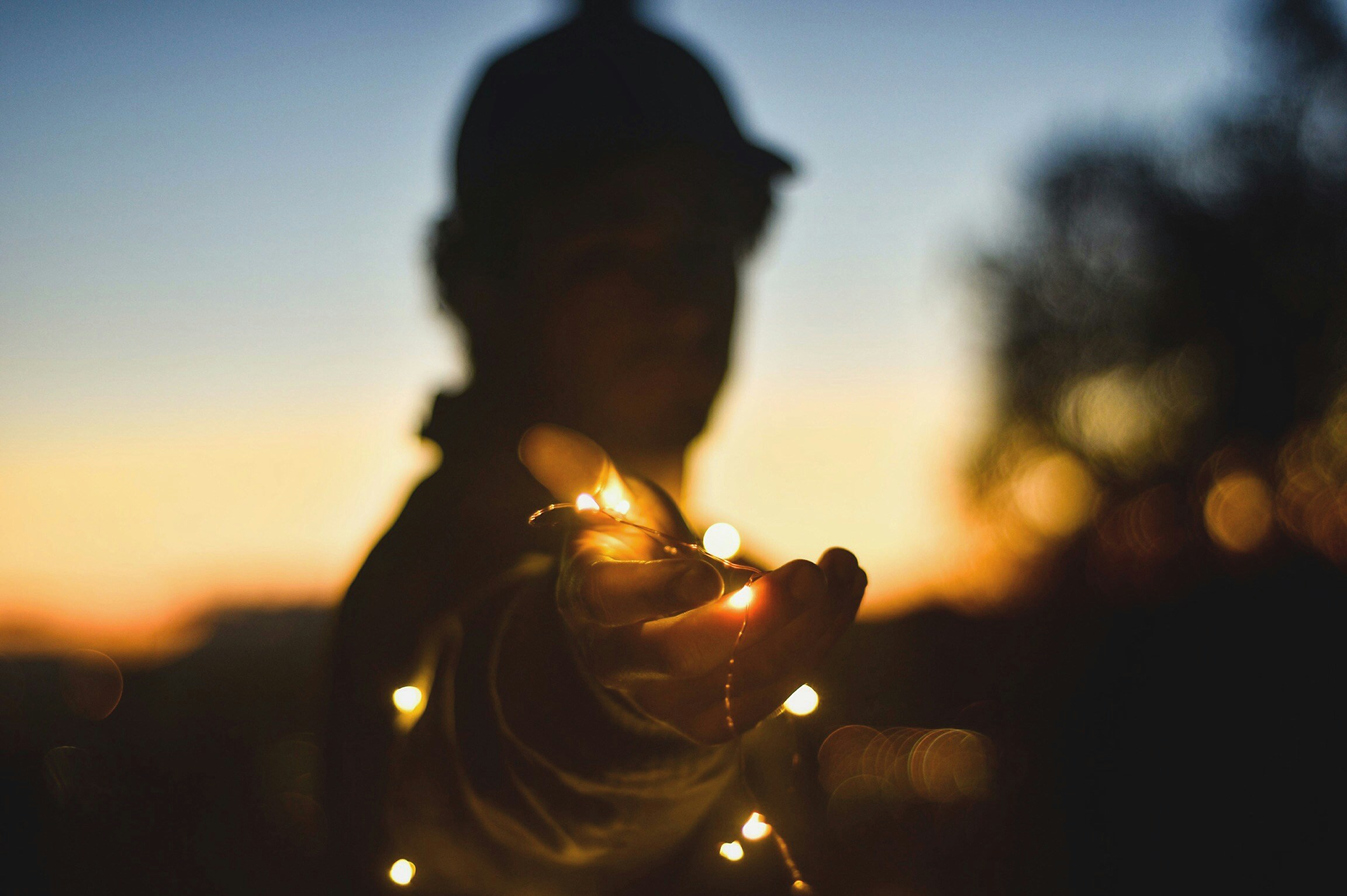 A backlit person at twilight holds a hand out towards the camera, holding some fairly lights