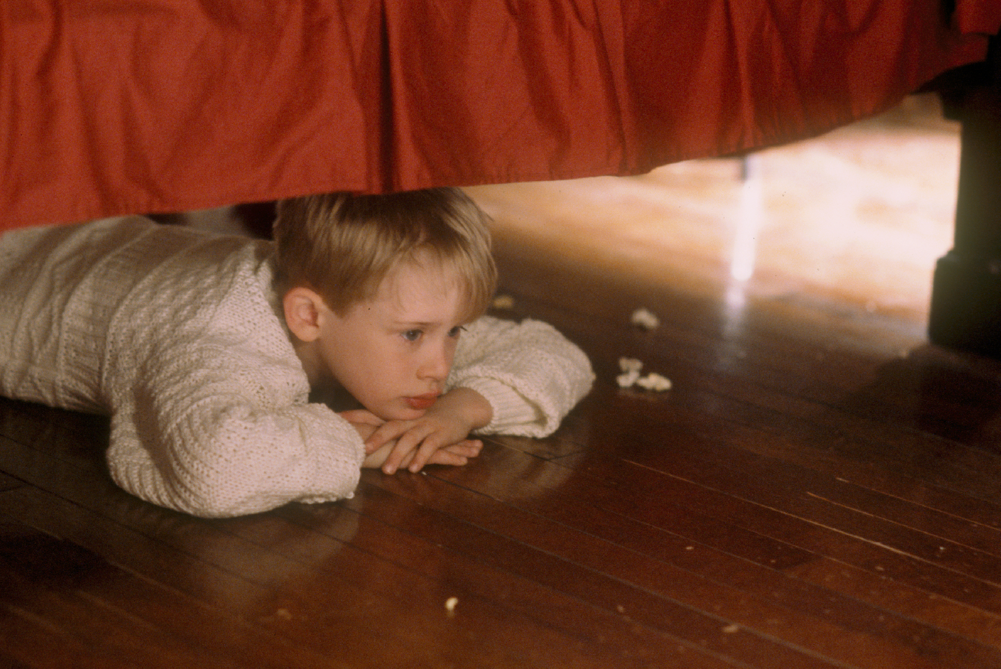 A child lies under a bed, resting his chin on his hands, contemplating.