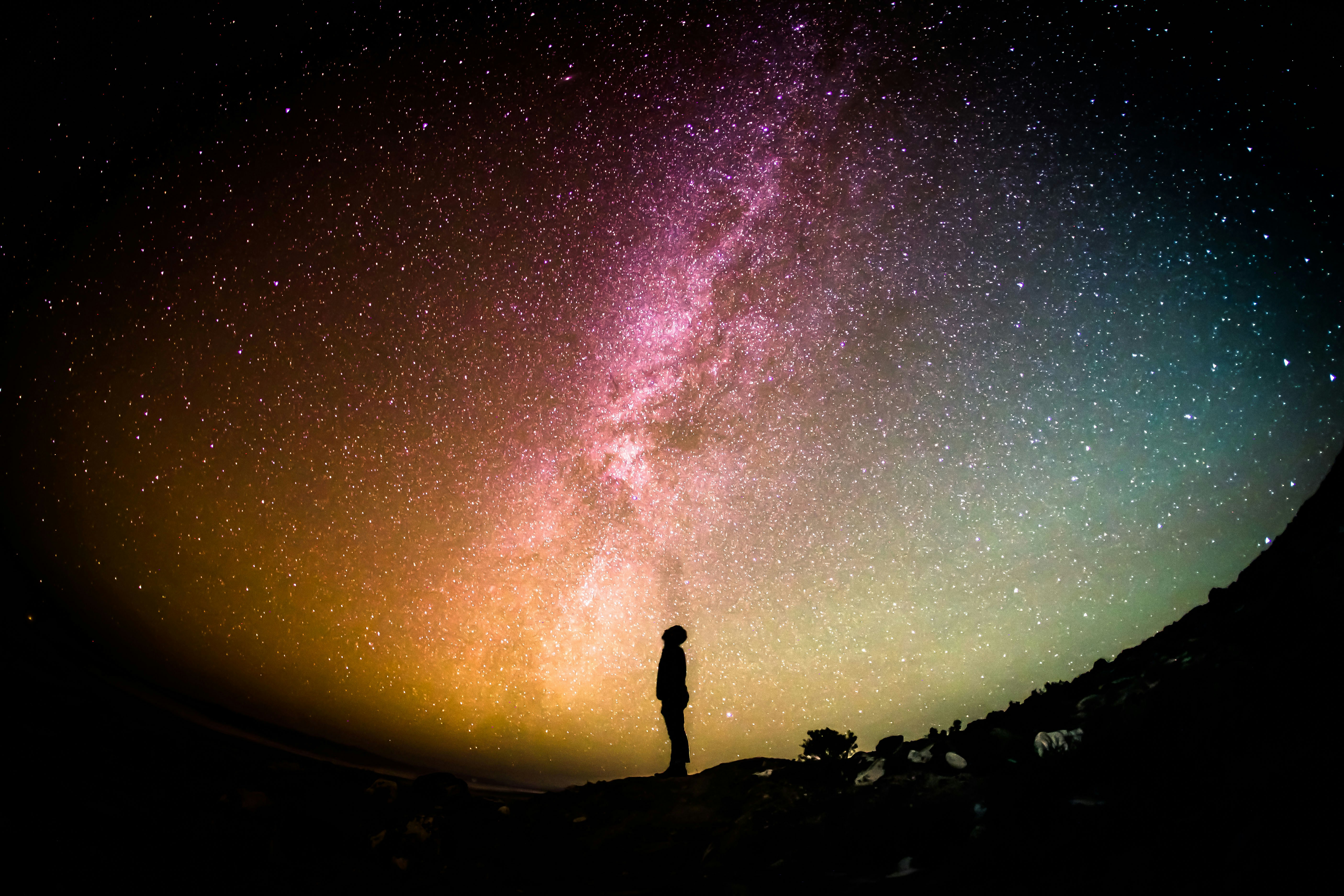 A fish eye lens view of a person standing silhouetted, looking up to a colourful night sky with the Milky Way across it