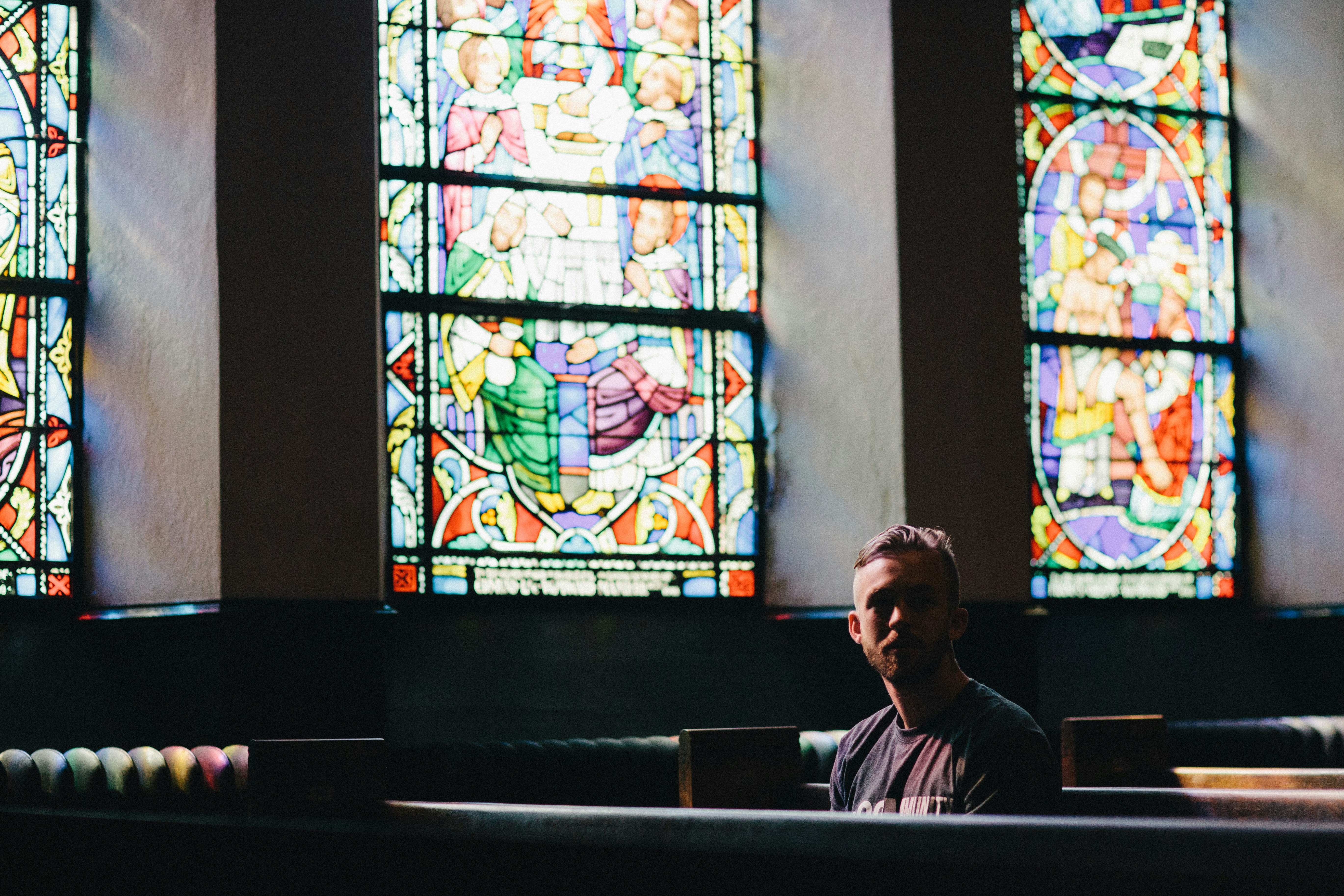 A man sits in a church pew below a colourful stained glass window, looking pensive.