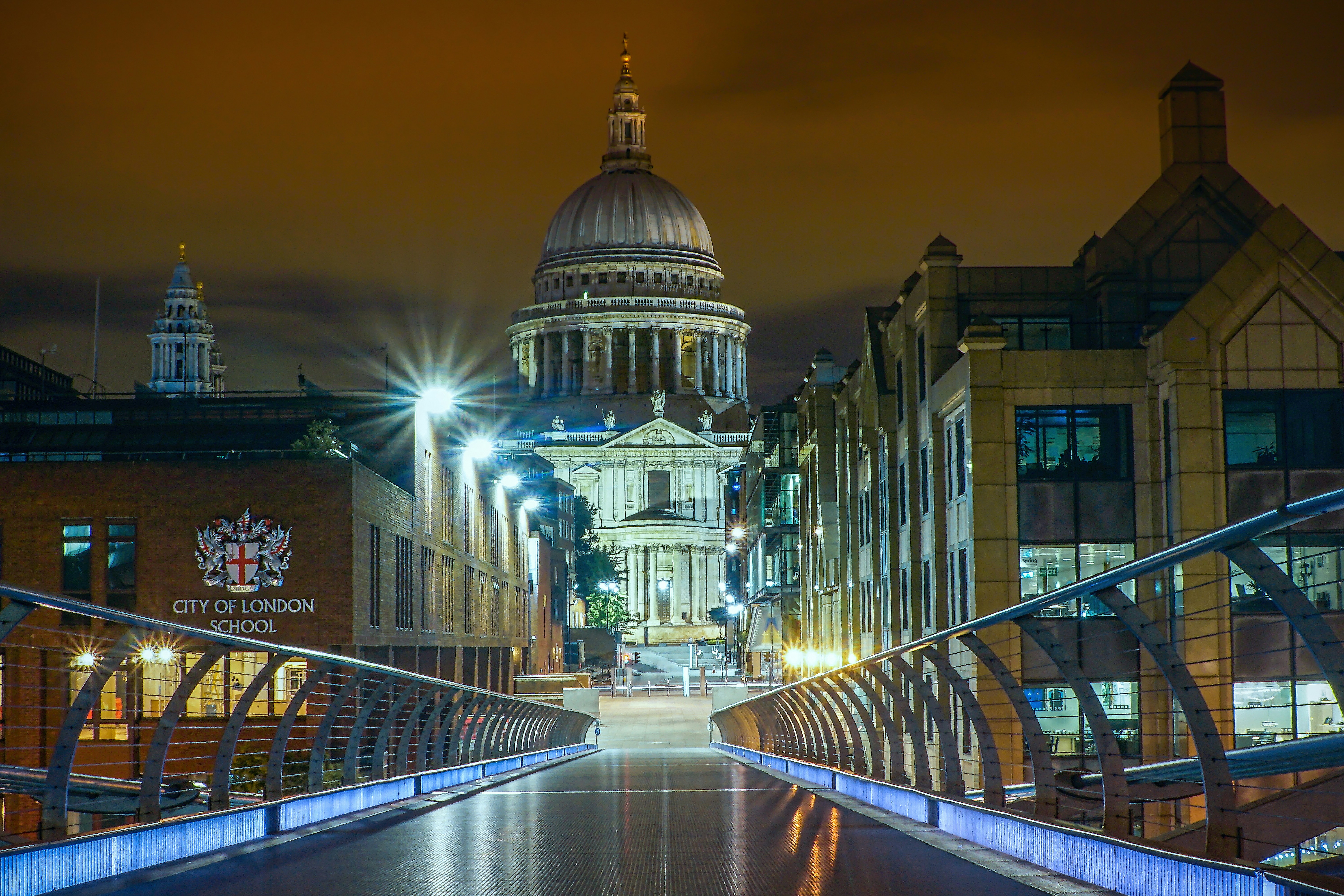 A view from a floodlight footbridge towards a gap between office buildings which reveal a cathedral and its illuminated dome.