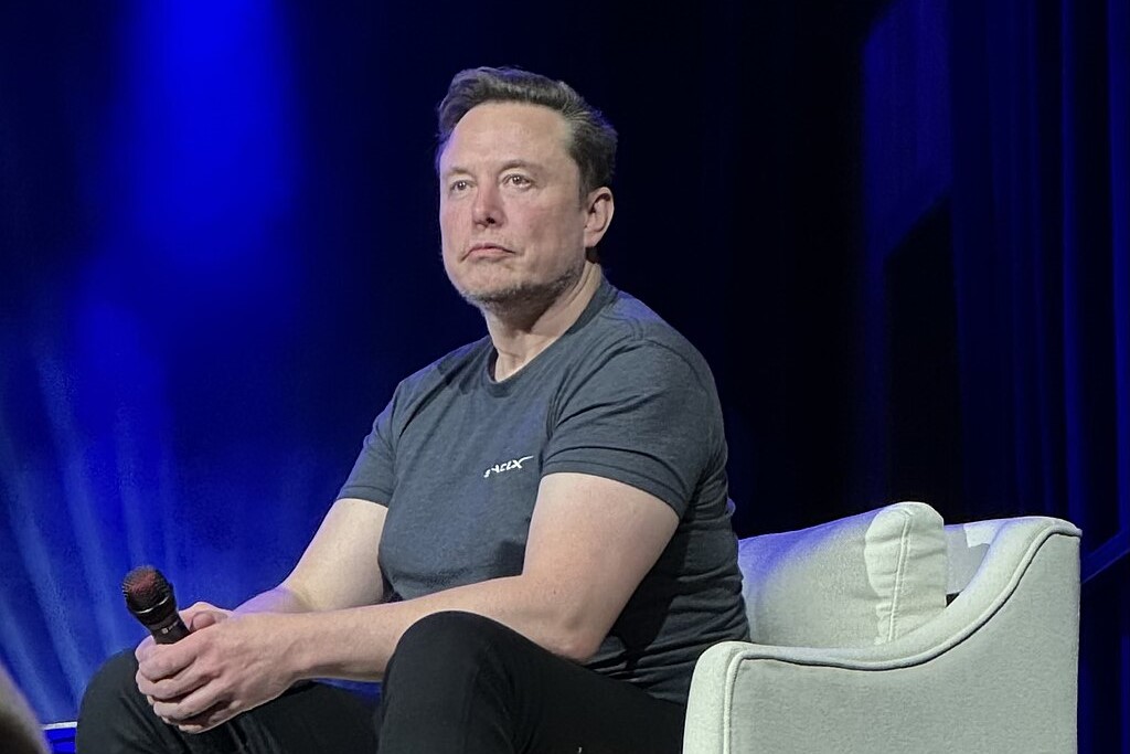 Elon Musk, wearing a t-shirt slouches forward, holding a mic, while sitting on a stage chair.