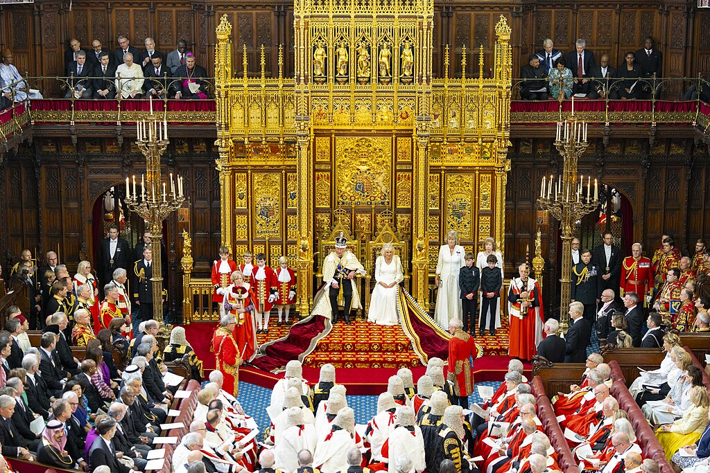 A wide angle picture shows a king and queen on thrones before many people in ceremonial clothes.