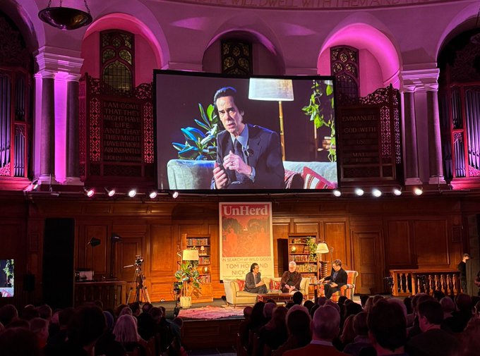 A view of an in-conversation on a stage, with a video screen above showing a close up.