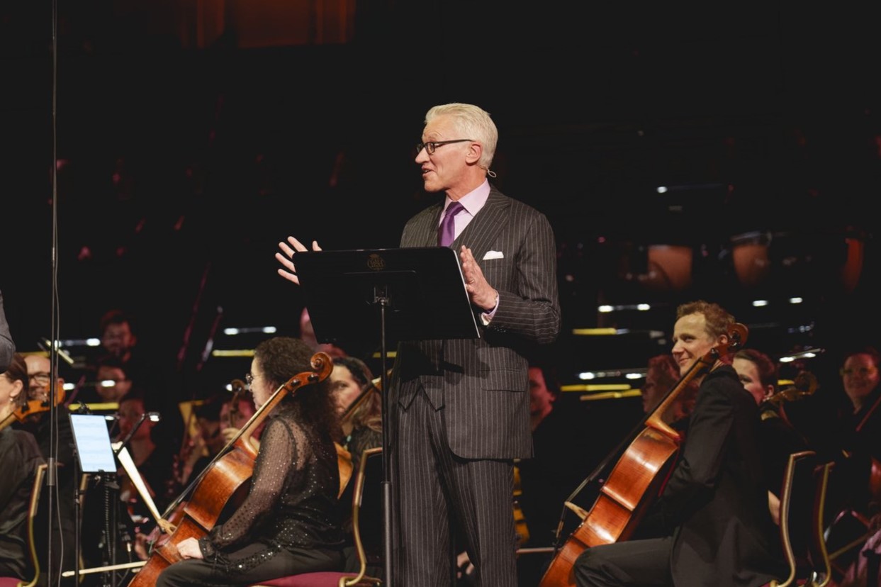 A man in a suit stands in front of a orchestra, by a lectern, gesturing while talking.