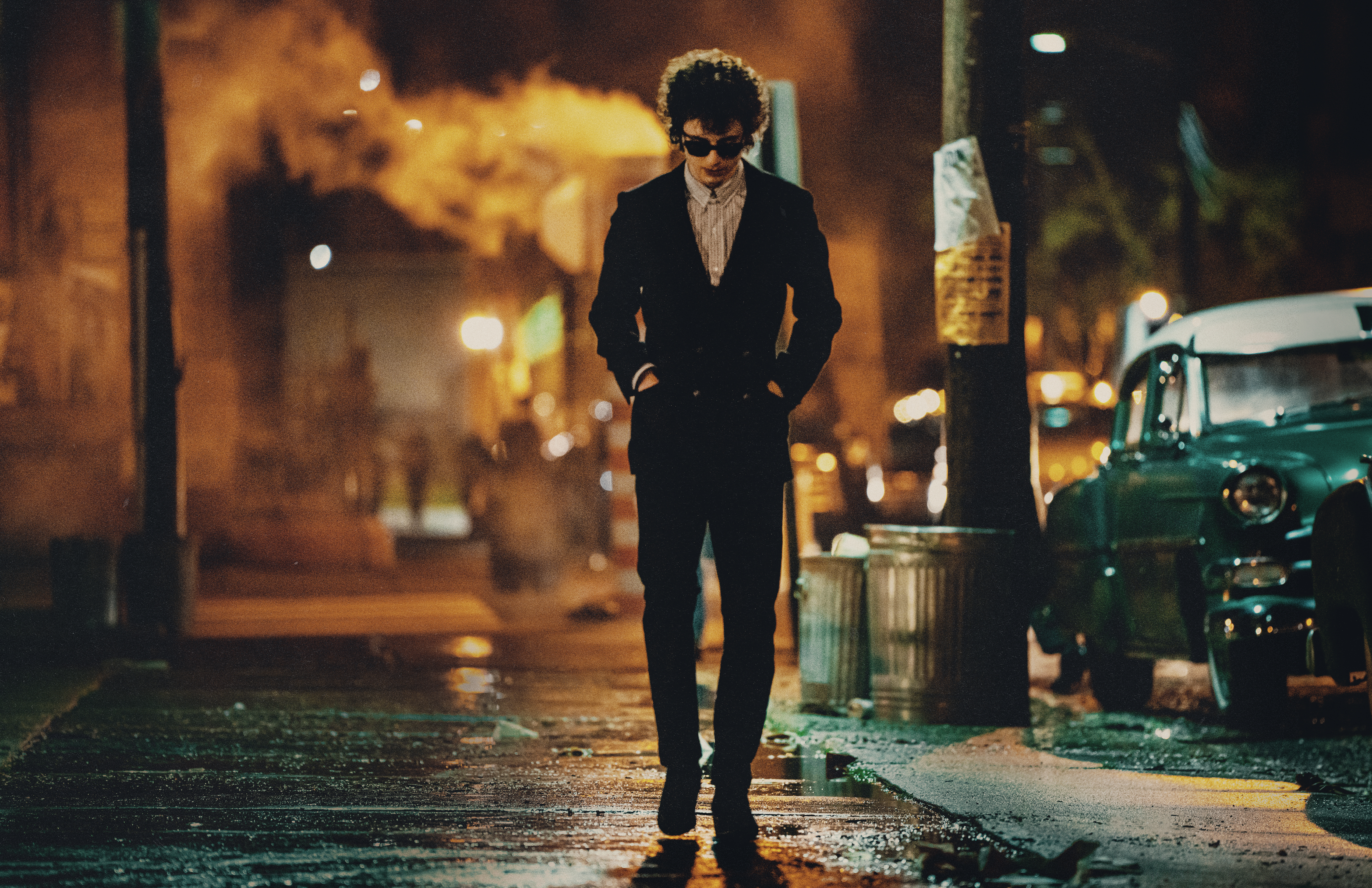 A hunched man wearing dark glasses and a dark suit, walks down a New York Street at night, with his hands in his pockets.