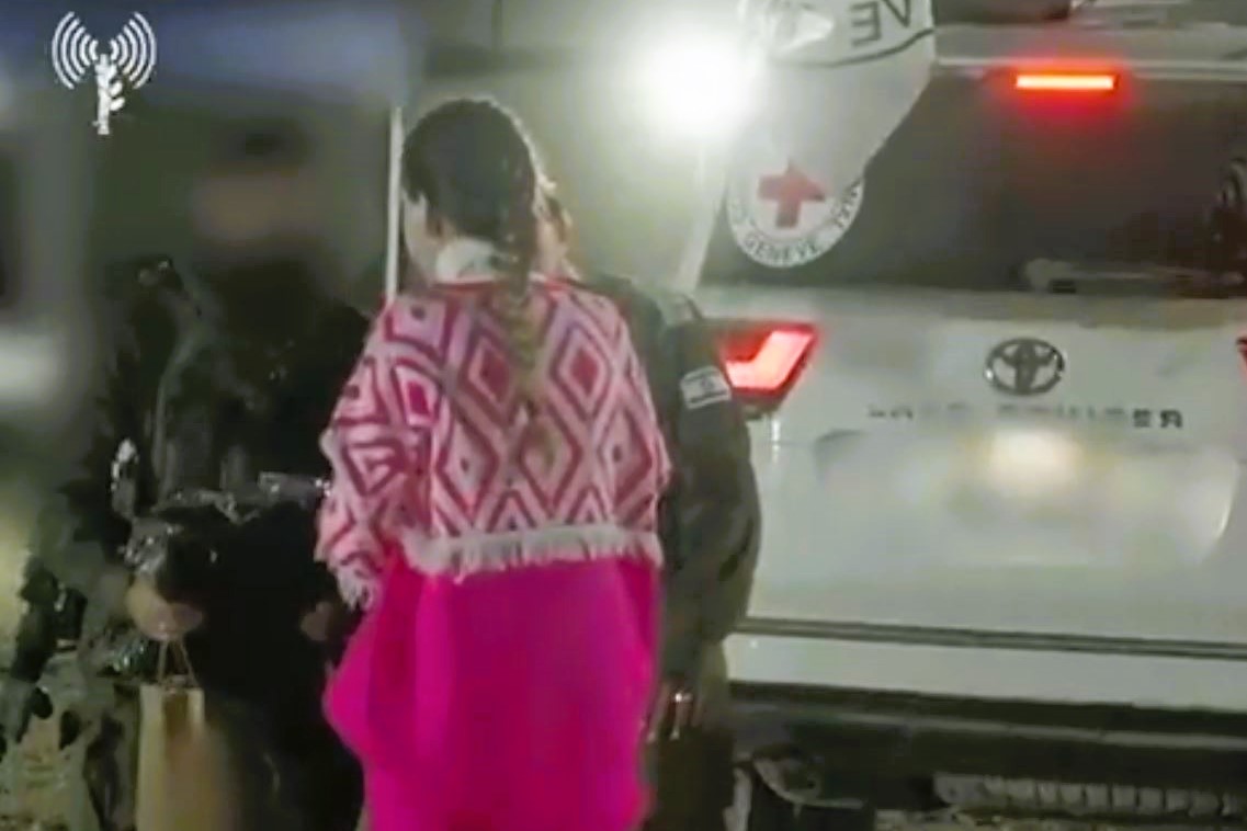 At a night time hostage exchange, a hostage wears a pink top and is guide toward the side of a white vehicle bearing a small red cross.