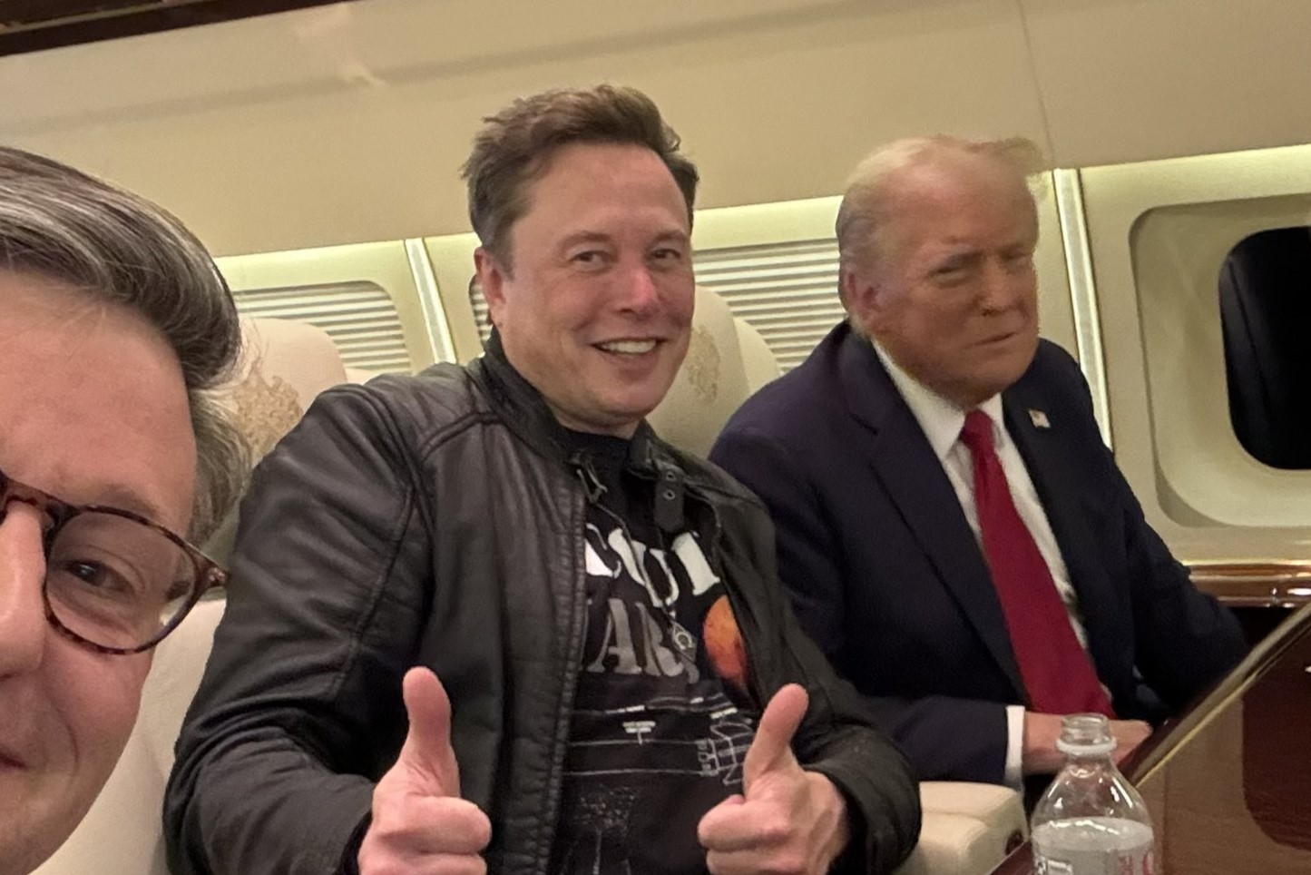 Elon Musk sits next to Donald Trump on a plan, while giving the thumbs up gesture with both hands.