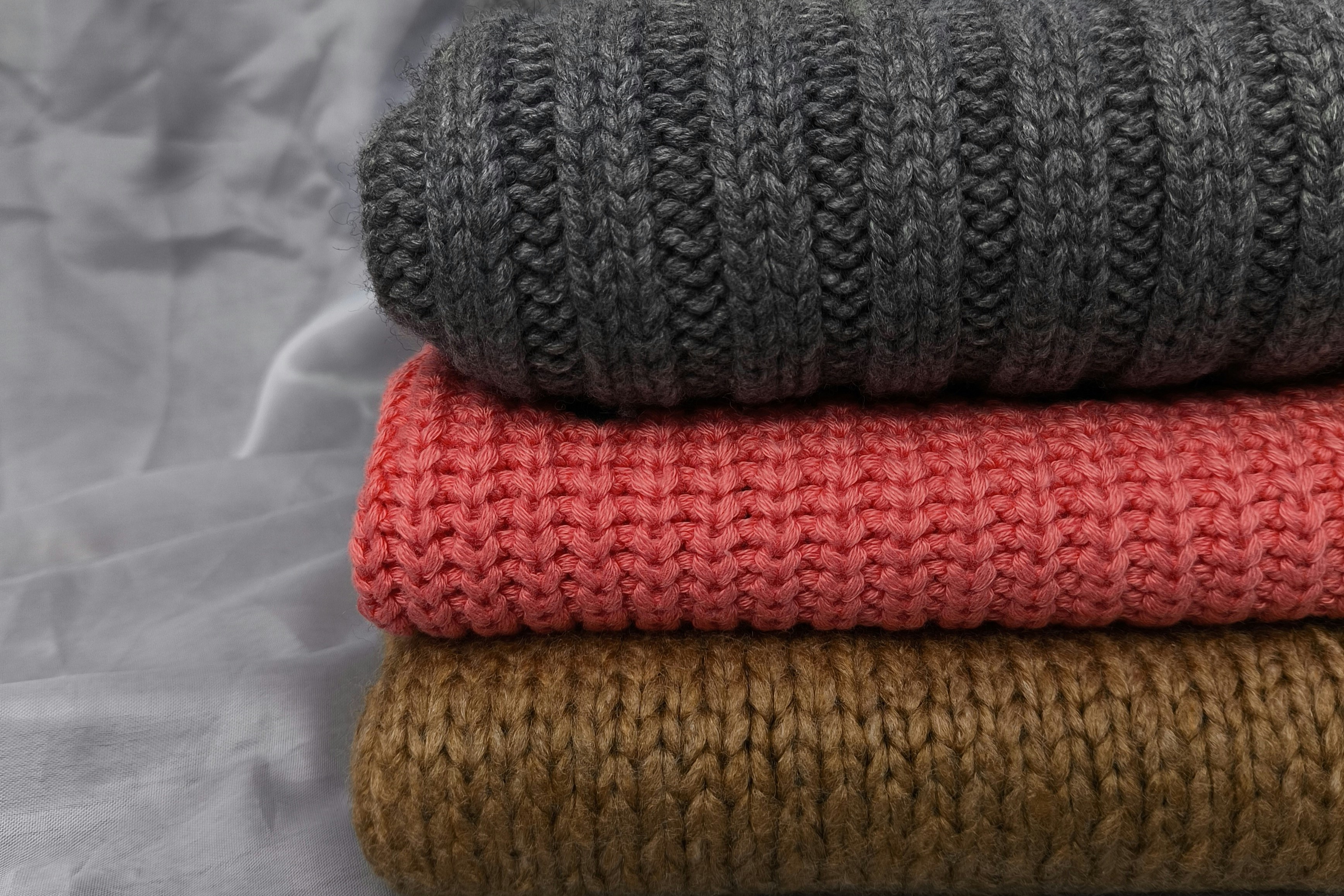 A pile of folded knitted jumpers sit on a grey back ground, one is also grey, the others are red and brown.