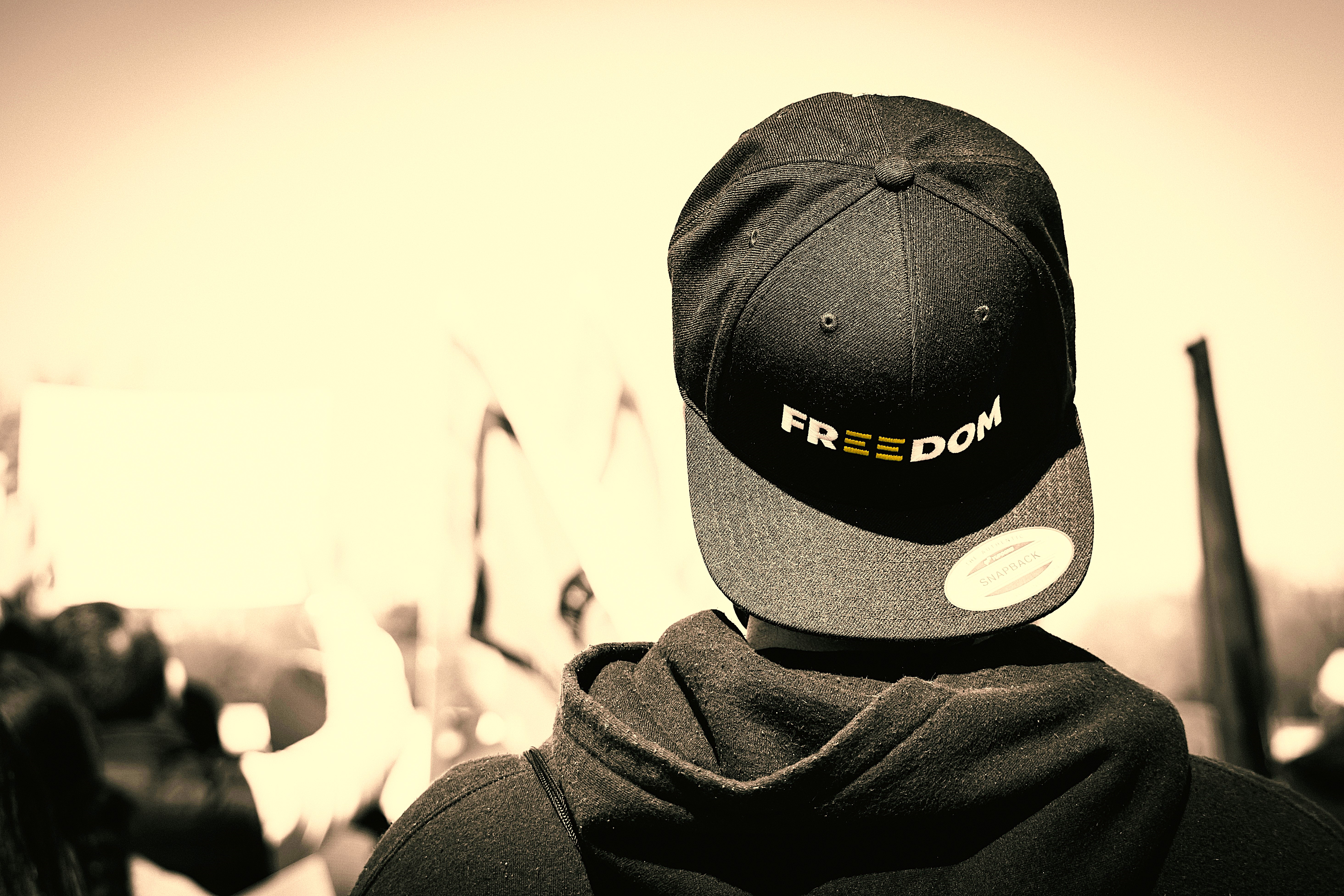 A protester stands with back to the camera, his baseball hat is turned backwards, it reads 'freedom'.