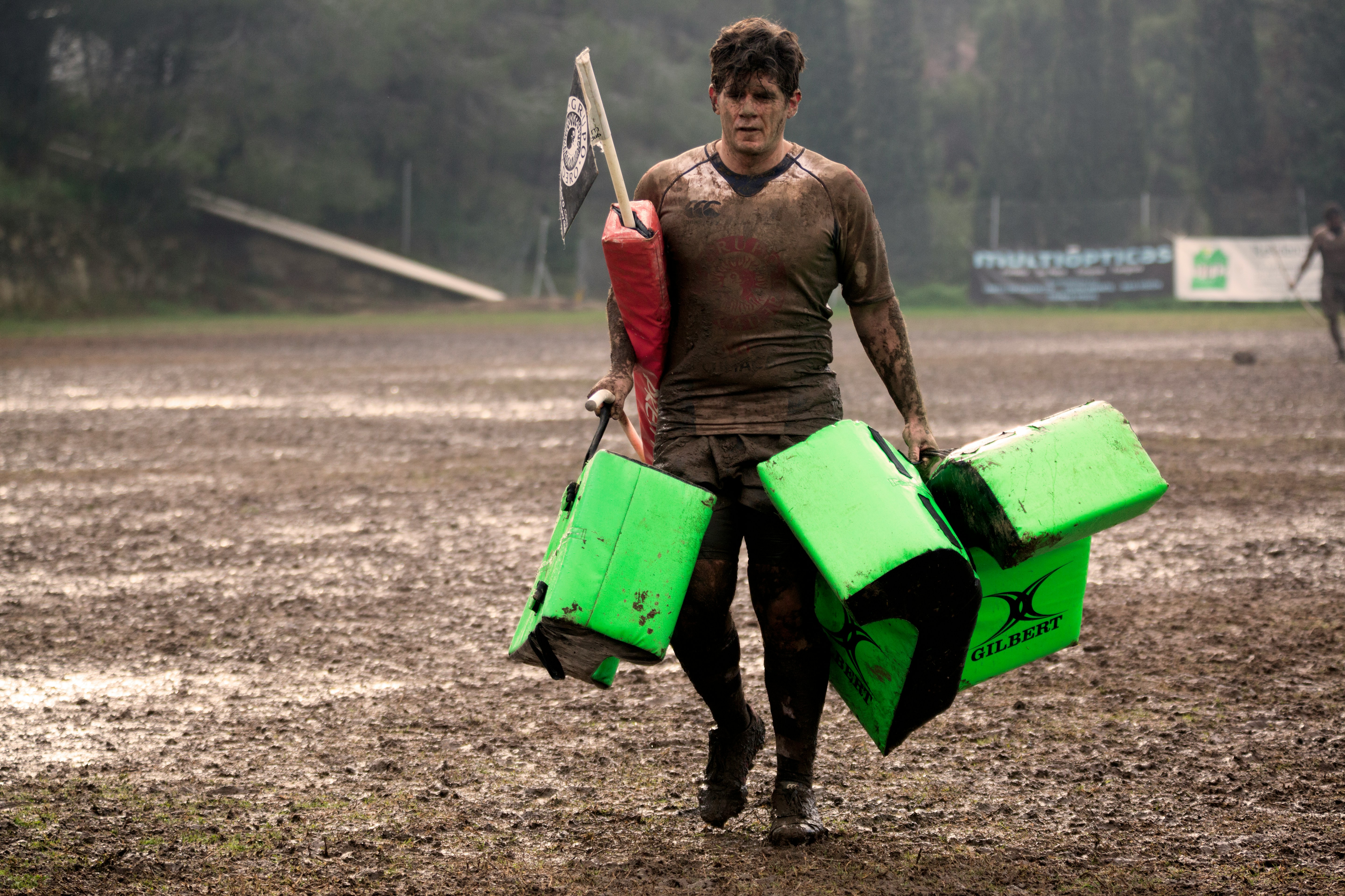 A muddy rugby player carries equipment off a pitch.