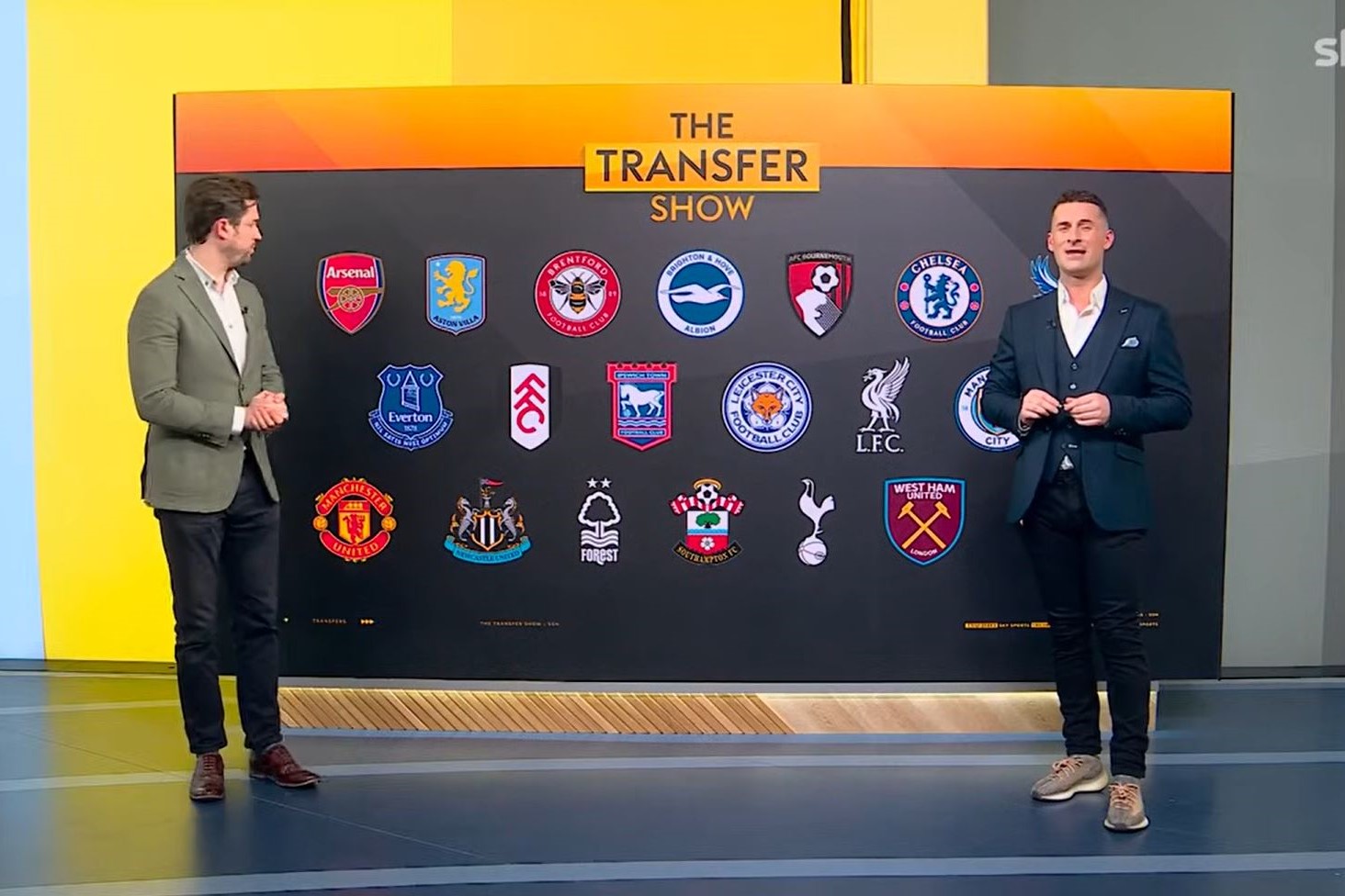 TV presenters stand eith side of a large screen showing football team badges.