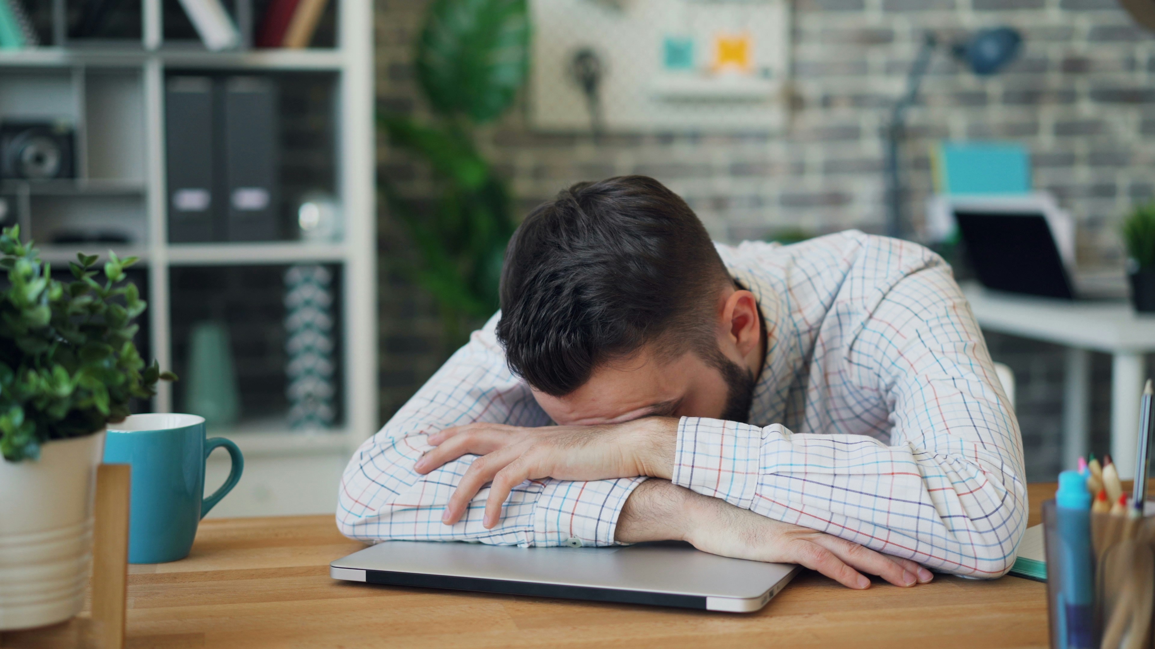 A man lies asleep on a closed lap top on a desk.