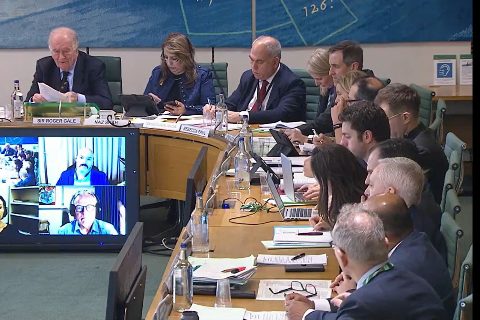 Members of a parliamentary committee sit at a curving table, in front of which a video screen shows other participants.