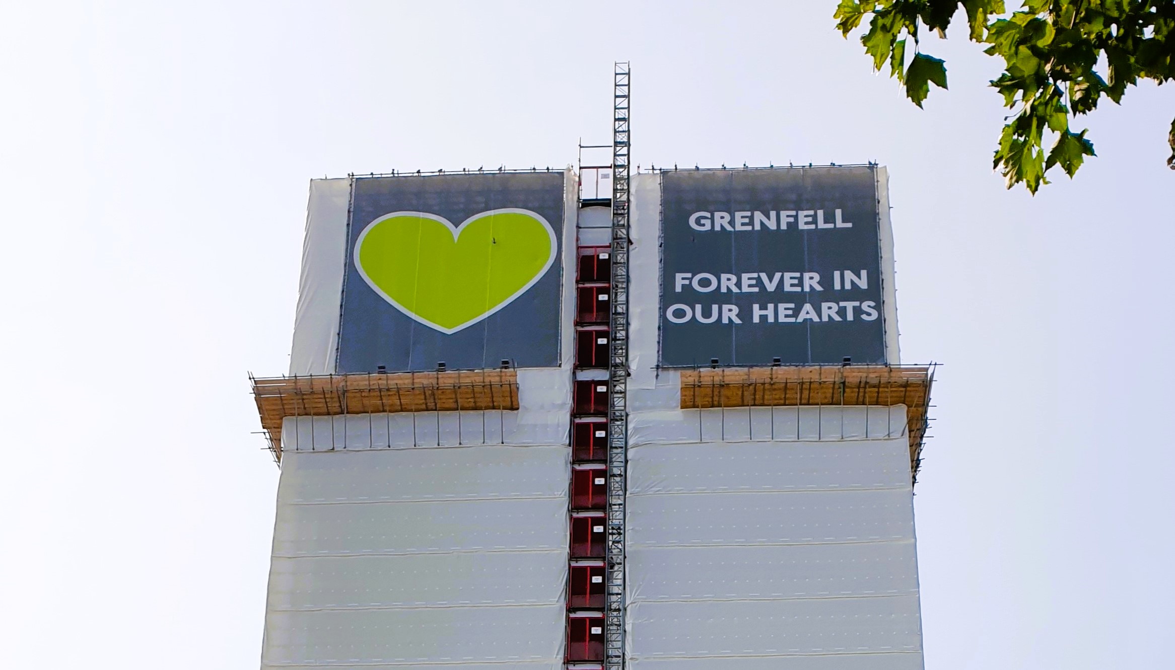 A white building wrap around a tower is topped by a green heart and the slogan: Grenfell Always In Our Heaets.