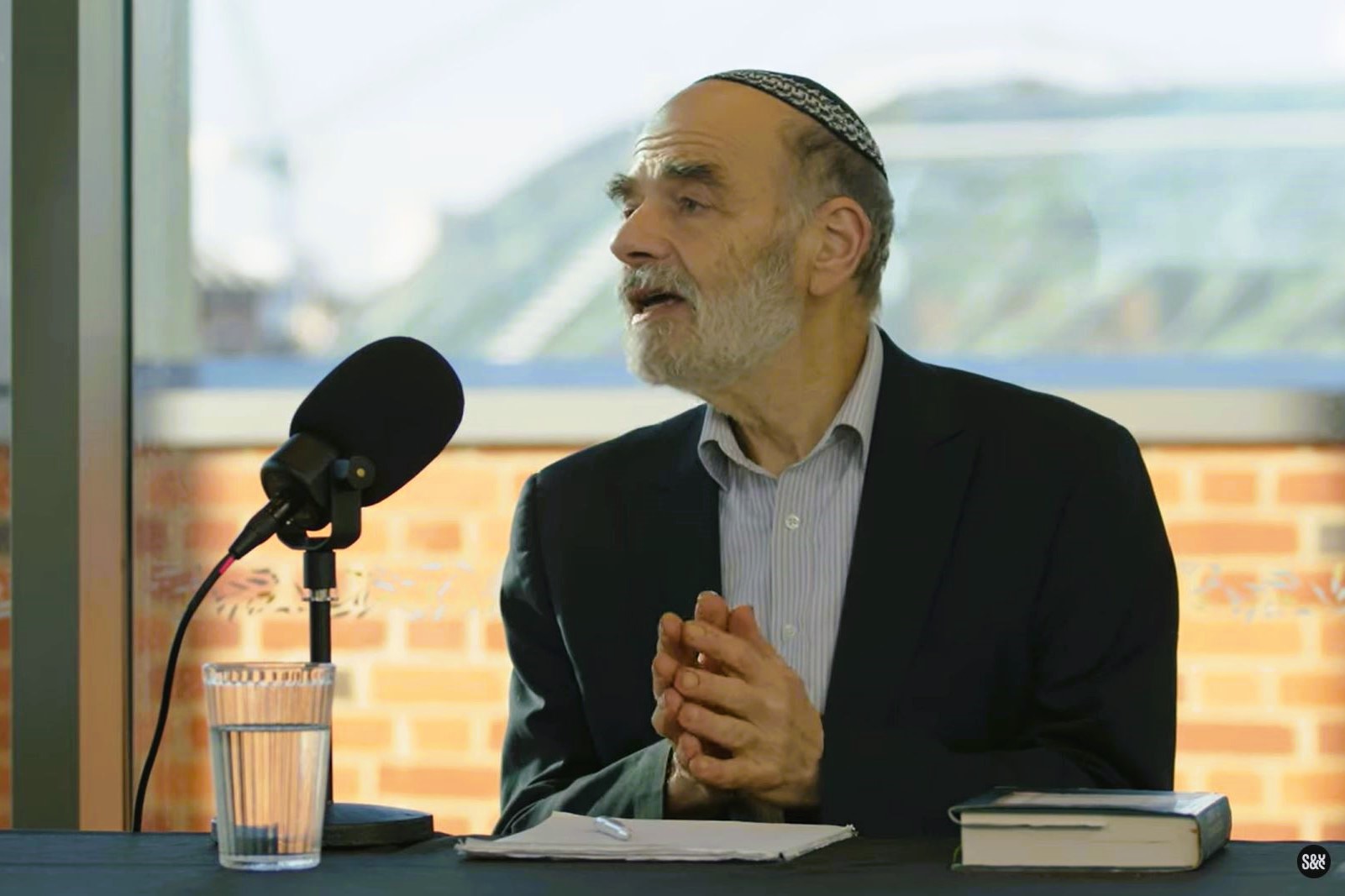 A podcast guest holds his hands together in front of himself as he raise his head in speech.