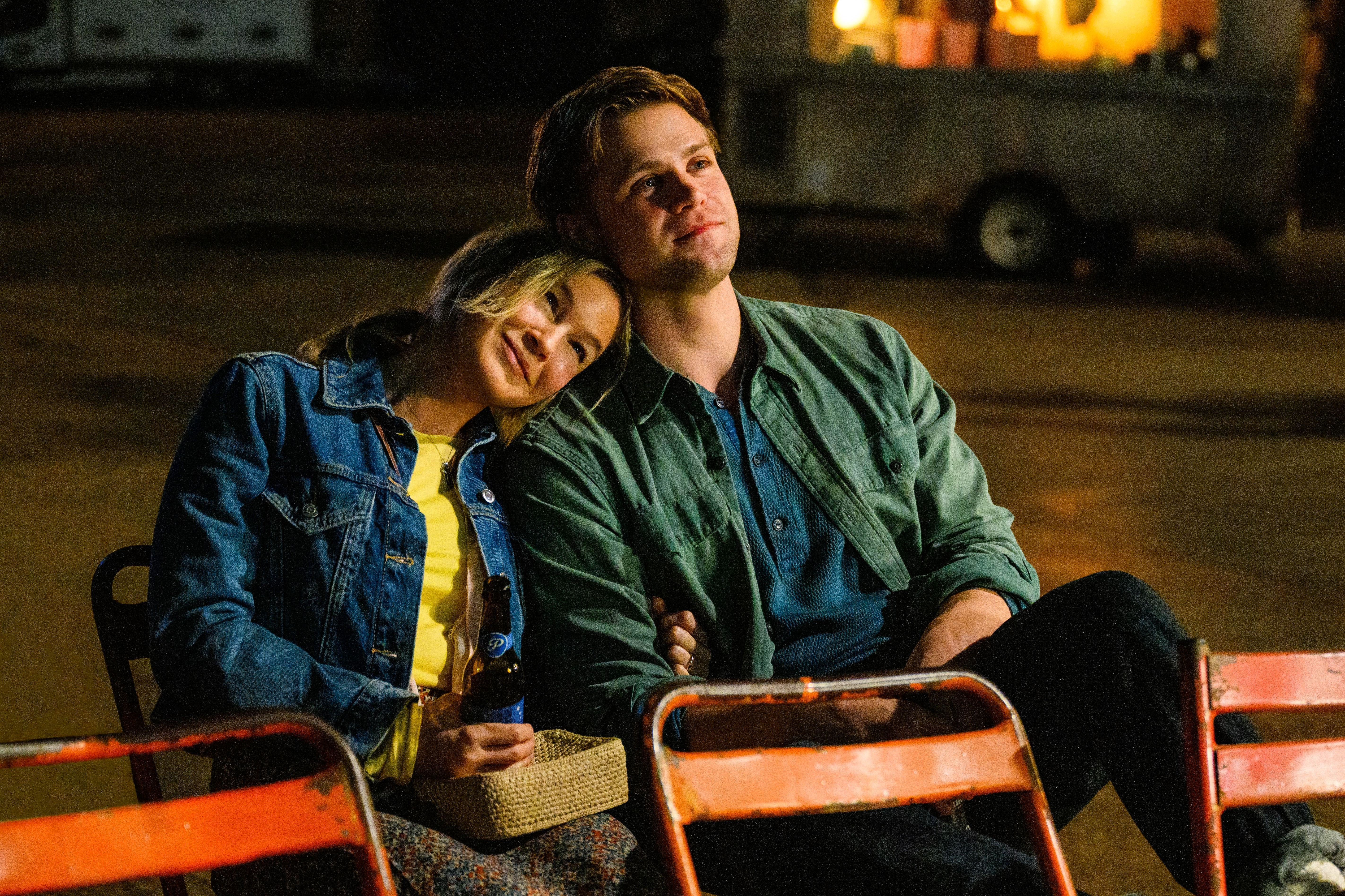 A couple sit on outdoor seats, her resting her head on his shoulder.