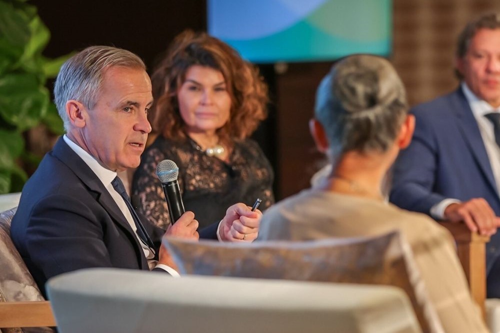 Mark Carney sits between two other speakers, holding a mic.