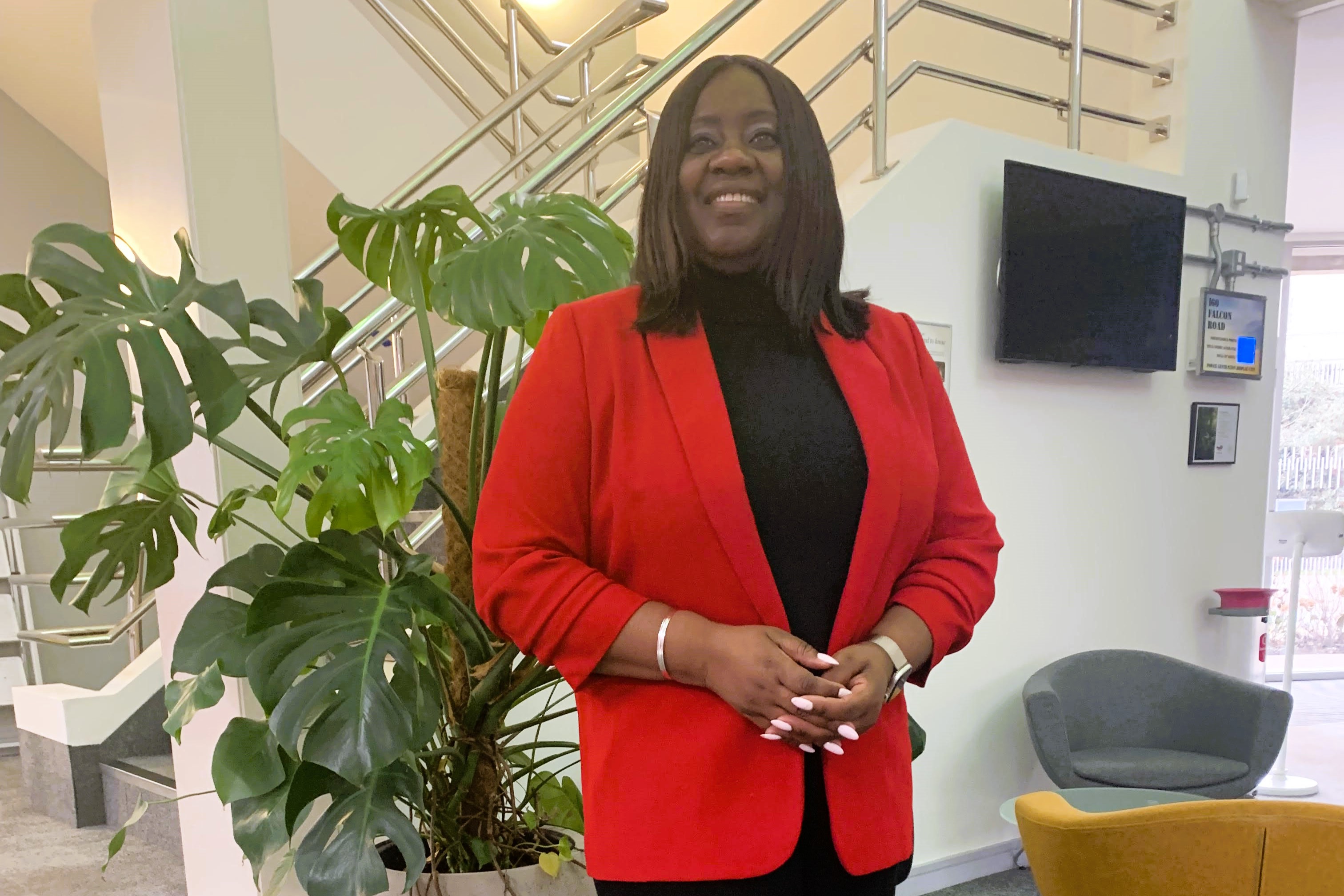 A woman wearing a red jacket stands formally beside an office stair case.