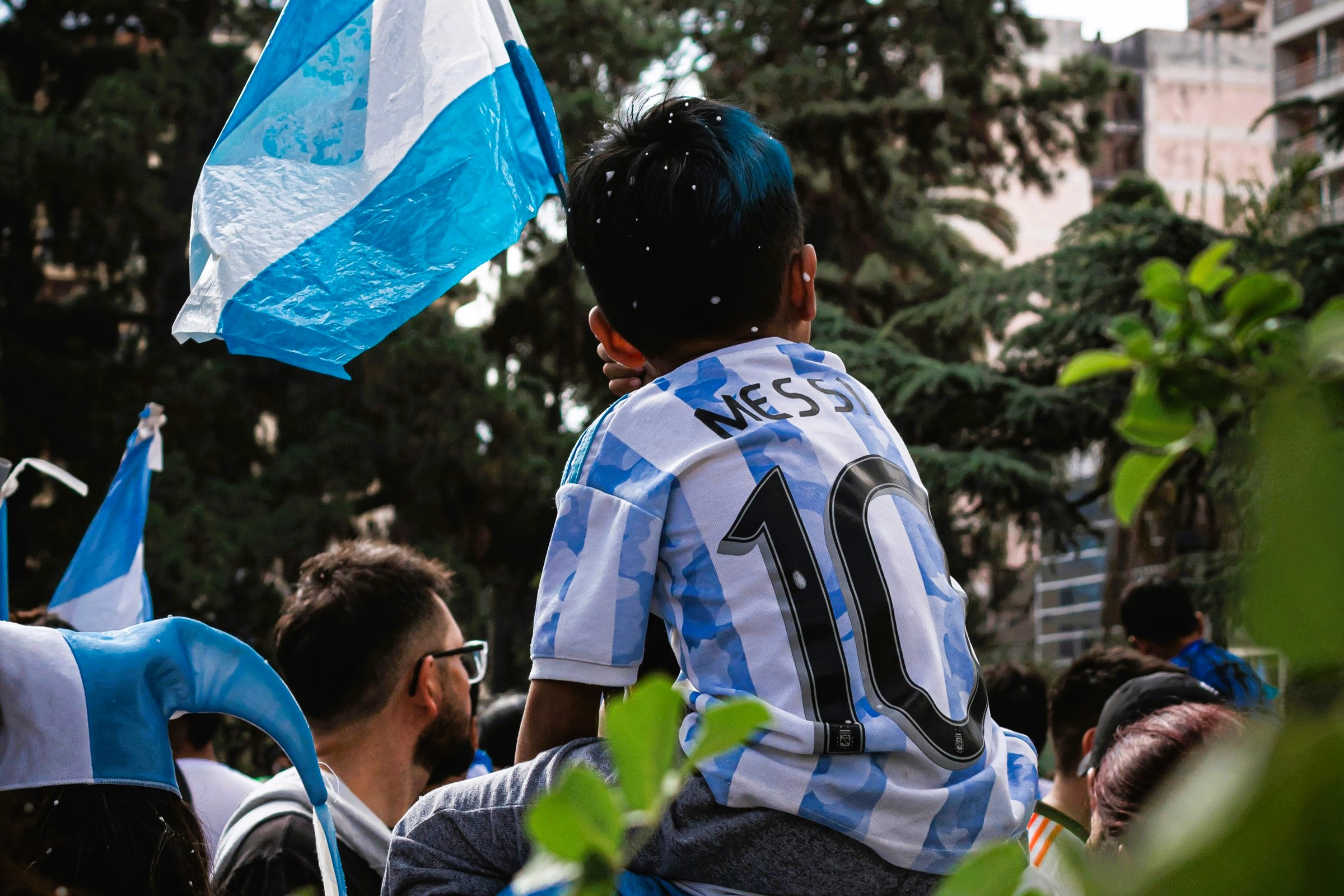 A child on the shoulders of a parent wears a light blue and white stripped football top, waves the Argentinian flag