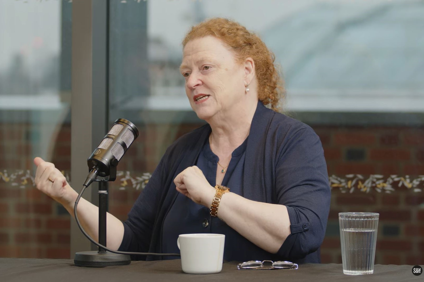 A woman guest on a podcast sits at a table with a mic in front, gesturing to the side with both hands.