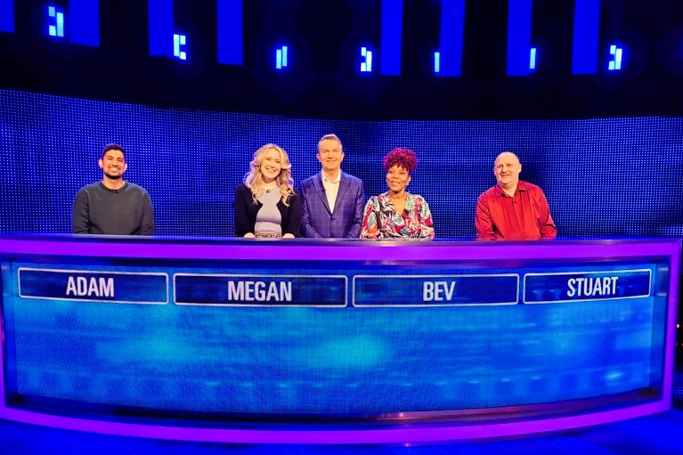 Four quiz show contestants stand behind a table with the show host.