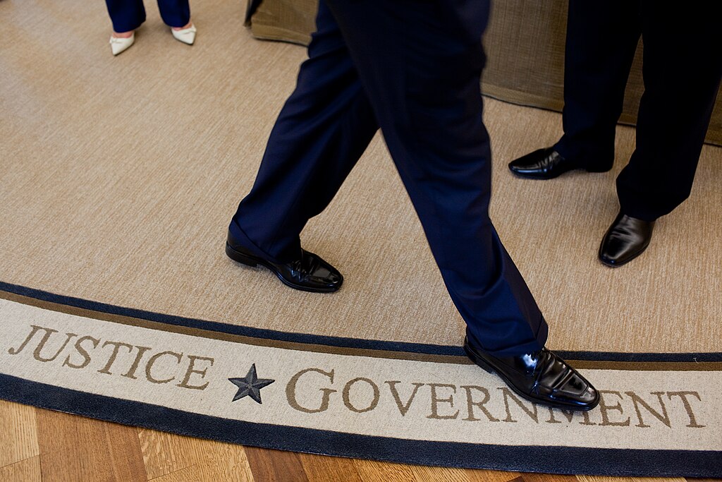 The feet and legs of someone walking on a white rug, beside the words Justice and Government woven in to it,