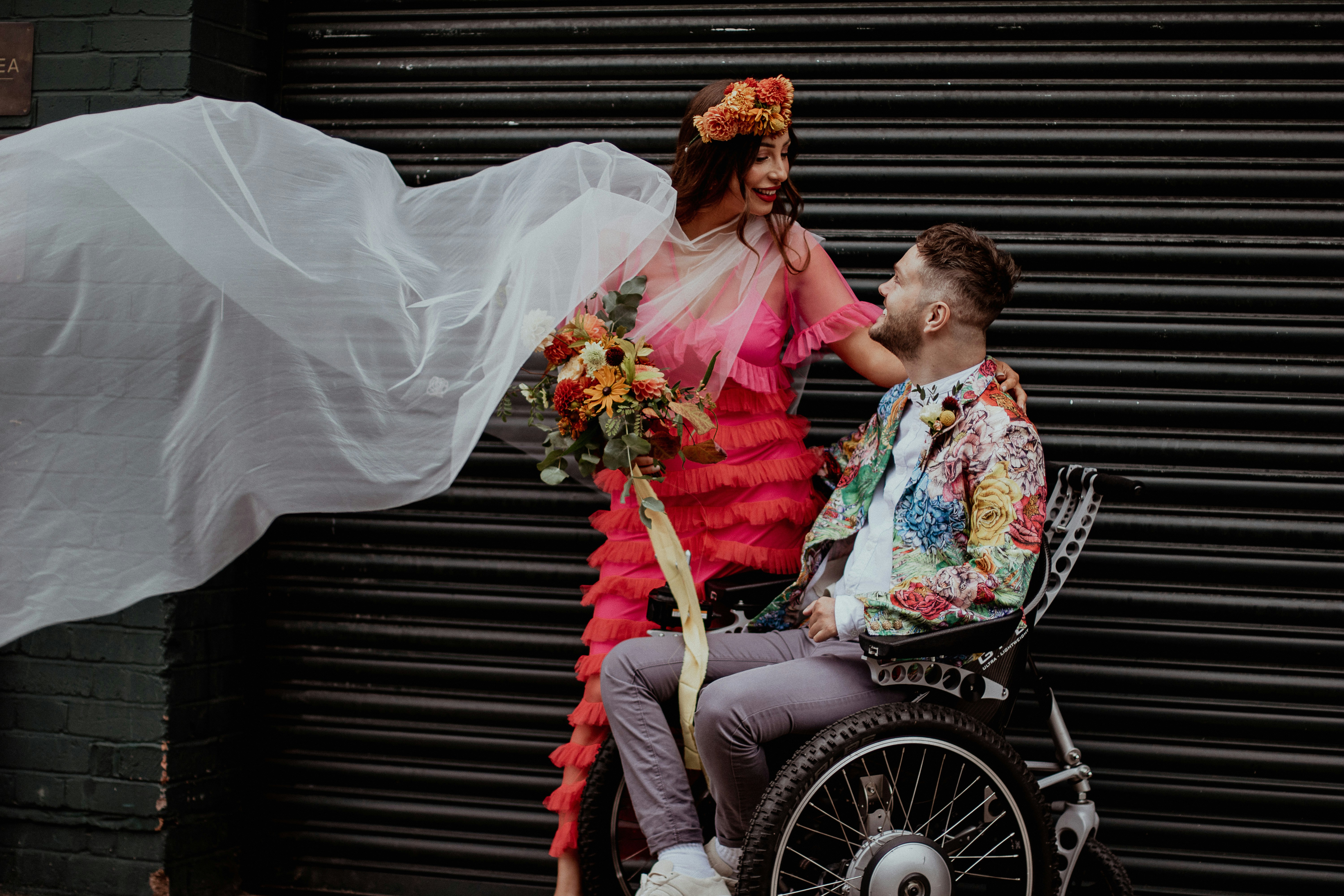 A bride dressed colourfully stands next to her groom, dressed similarly, as he sits in a wheelchair.