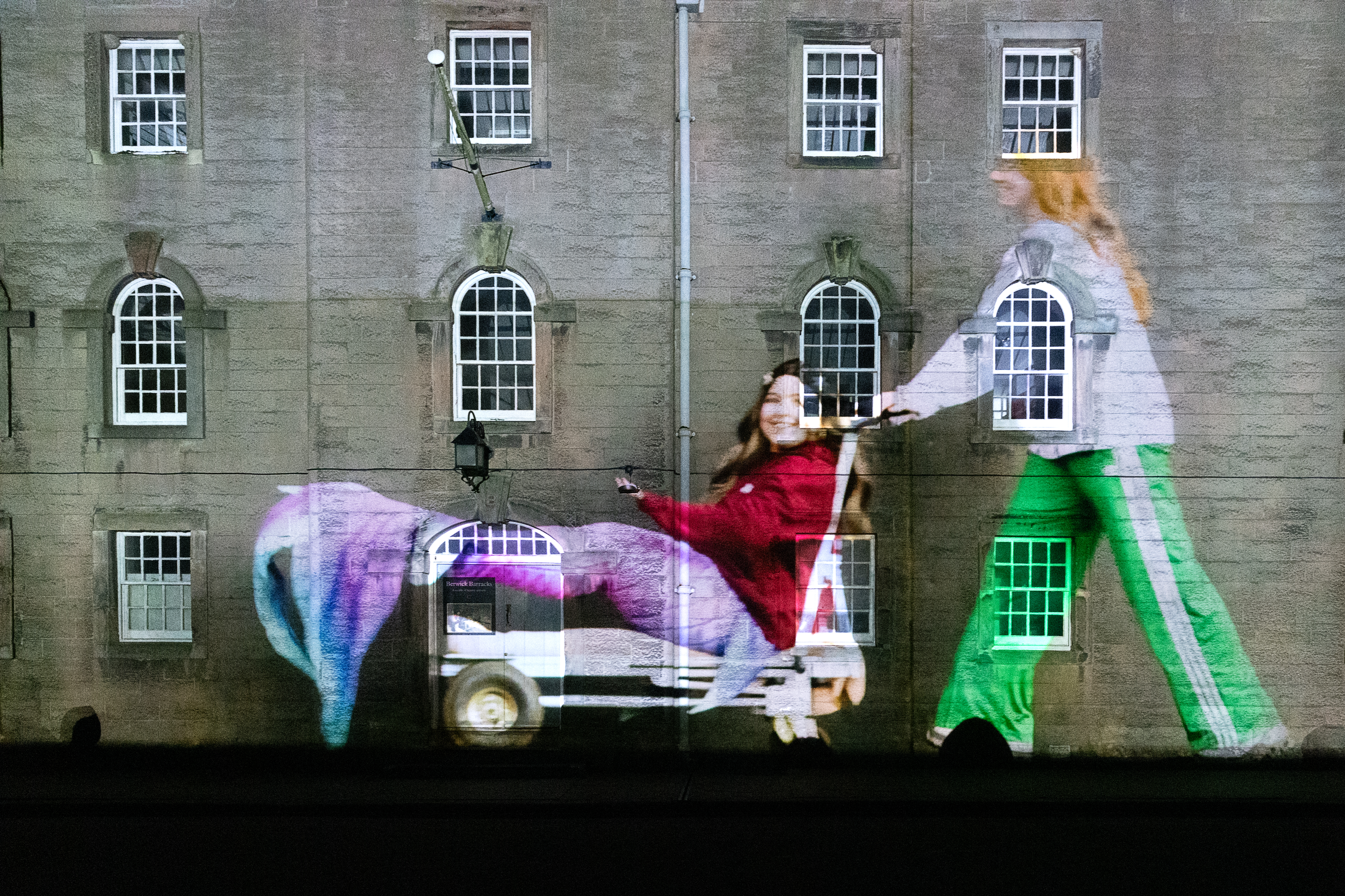 A projected image of a person dressed as a mermaid being pushed on a trolley falls on the wall and windows of an old barracks.