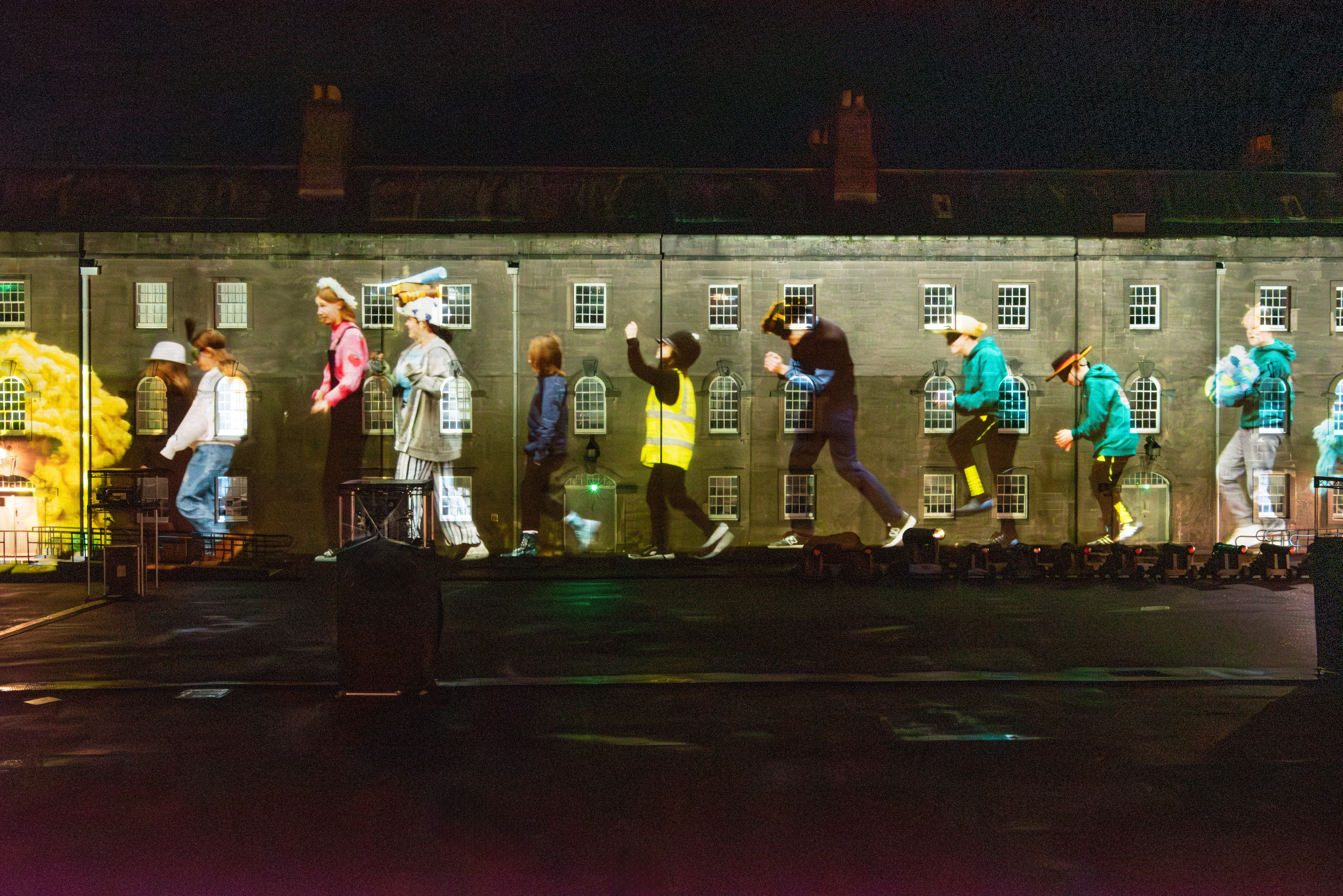 A projection of a parade of young people dressed theatrically against a barracks wall.